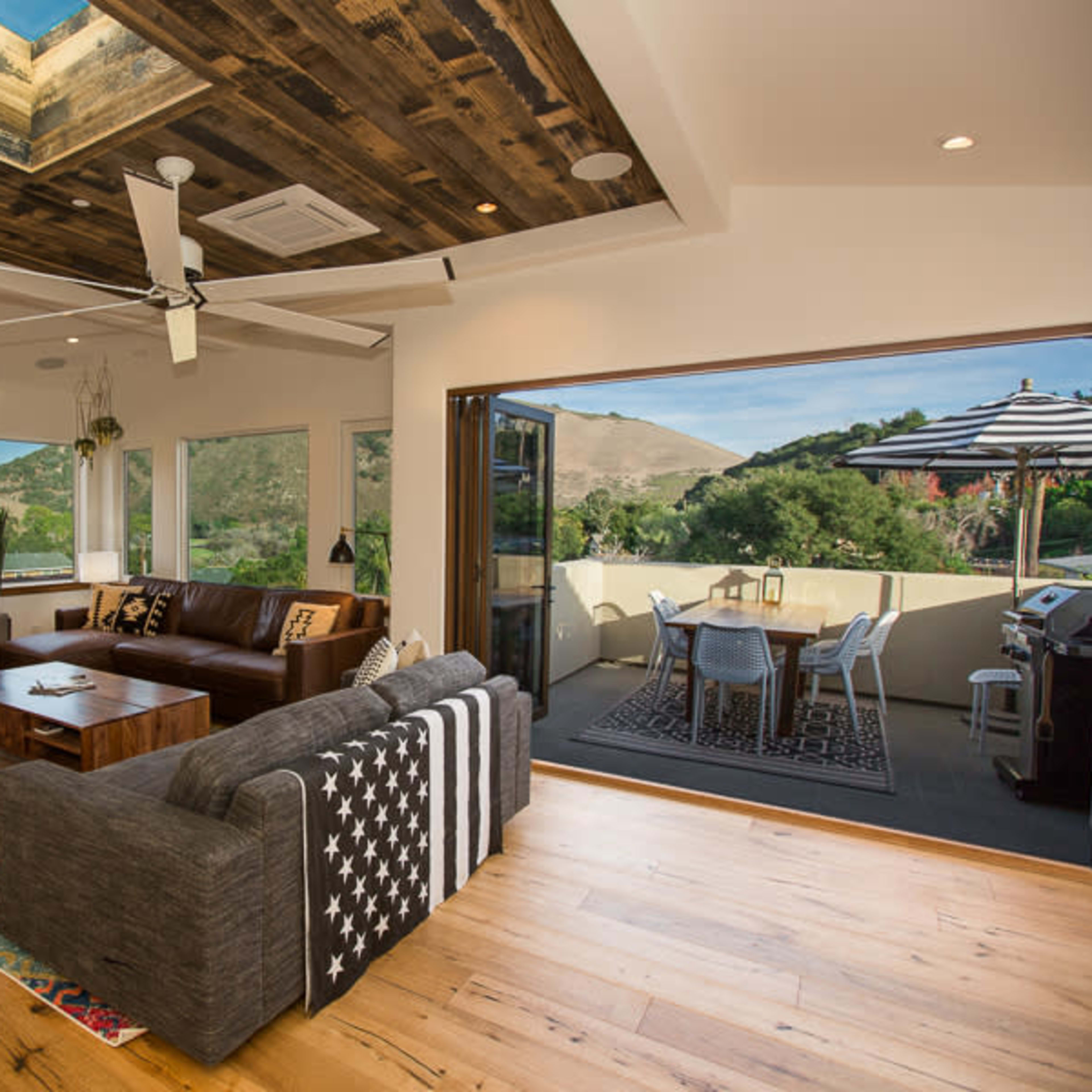 A modern living room with a seating area, a television above a fireplace, and large doors leading to a patio with outdoor dining furniture.