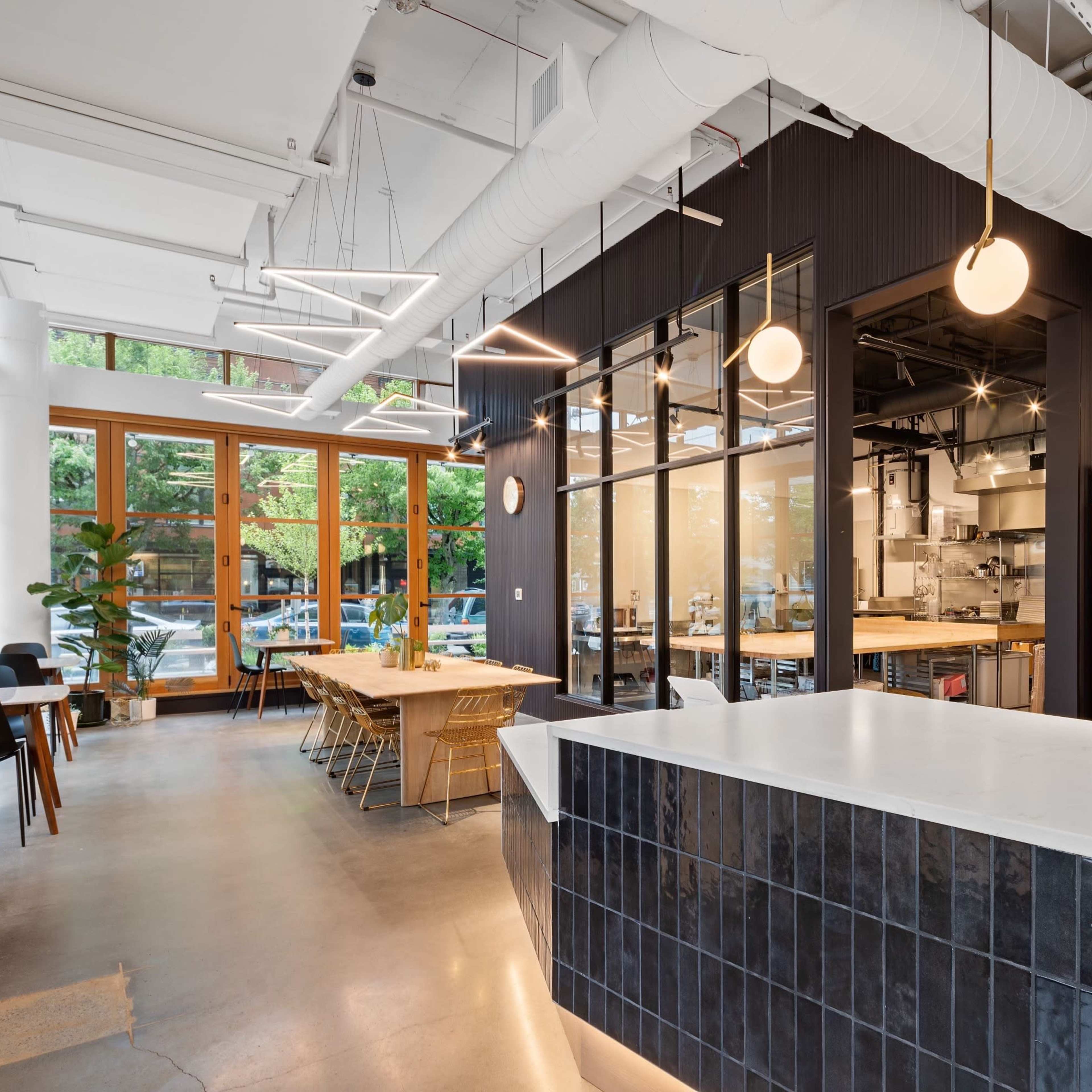 A modern cafe interior with large windows, a long white countertop, wooden tables, and a visible kitchen area in the background.