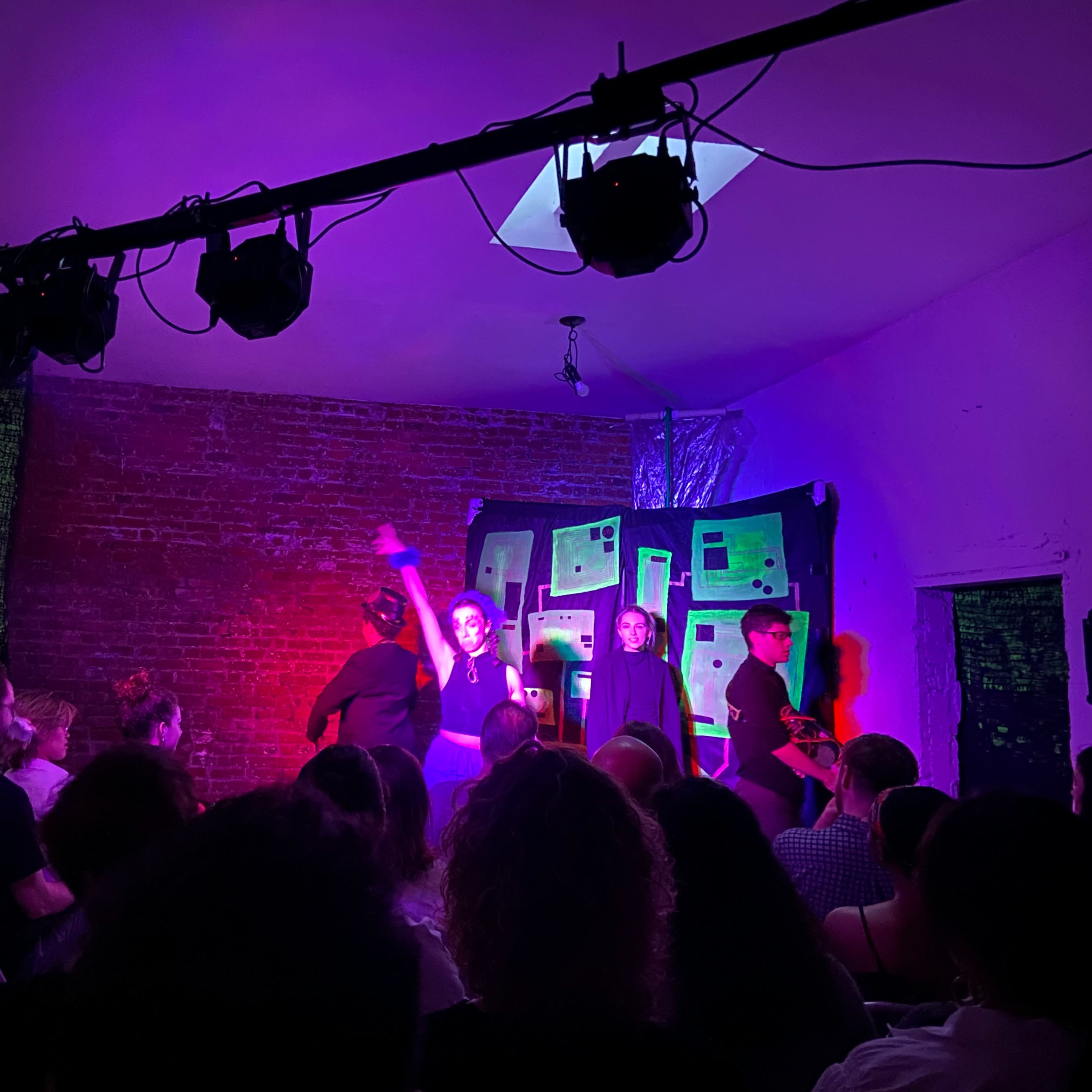 A group of performers is on stage during a theatrical performance, while an audience observes from their seats in a dimly lit room.