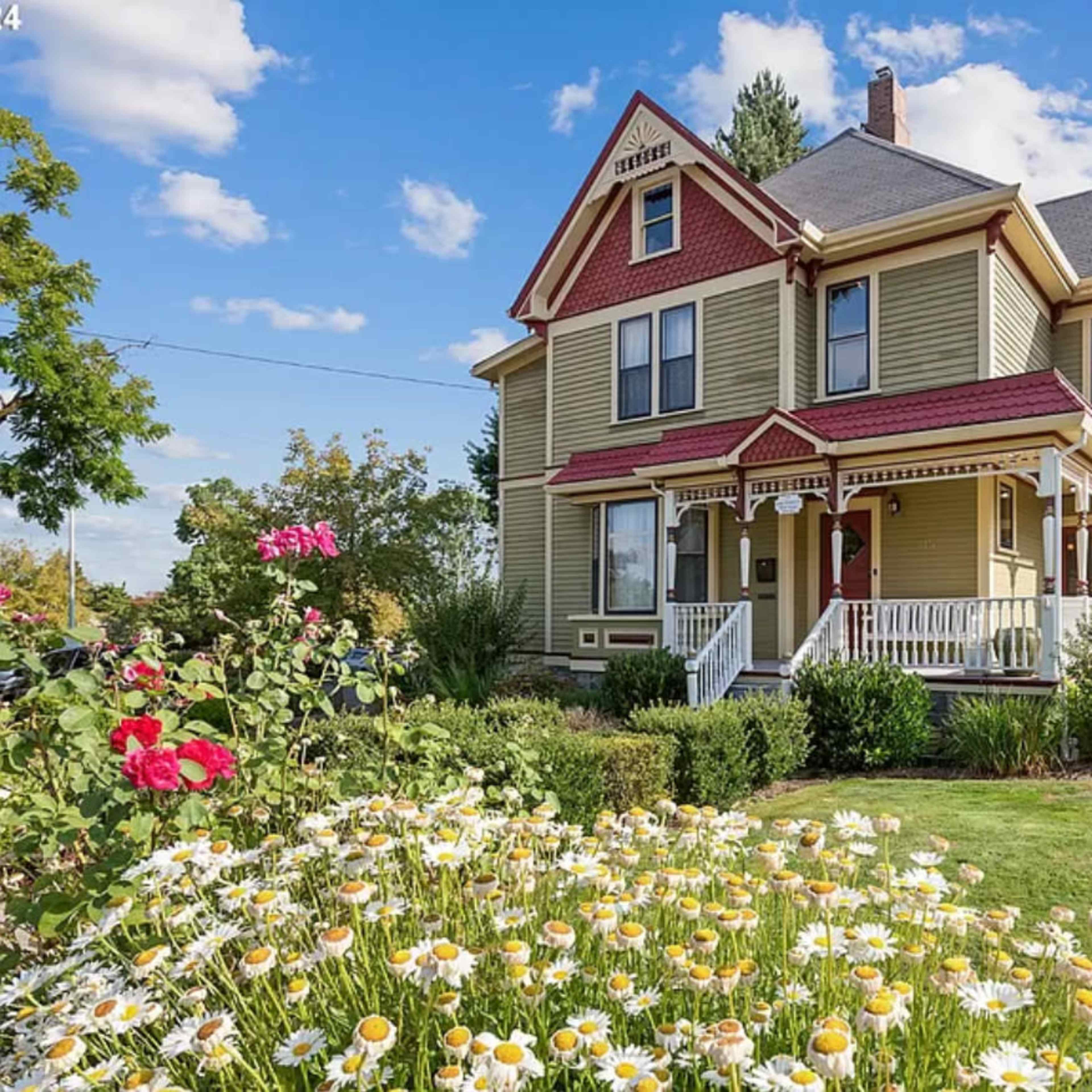 A Victorian-style house with a green exterior and red accents is surrounded by a garden of daisies and roses under a blue sky with scattered clouds.