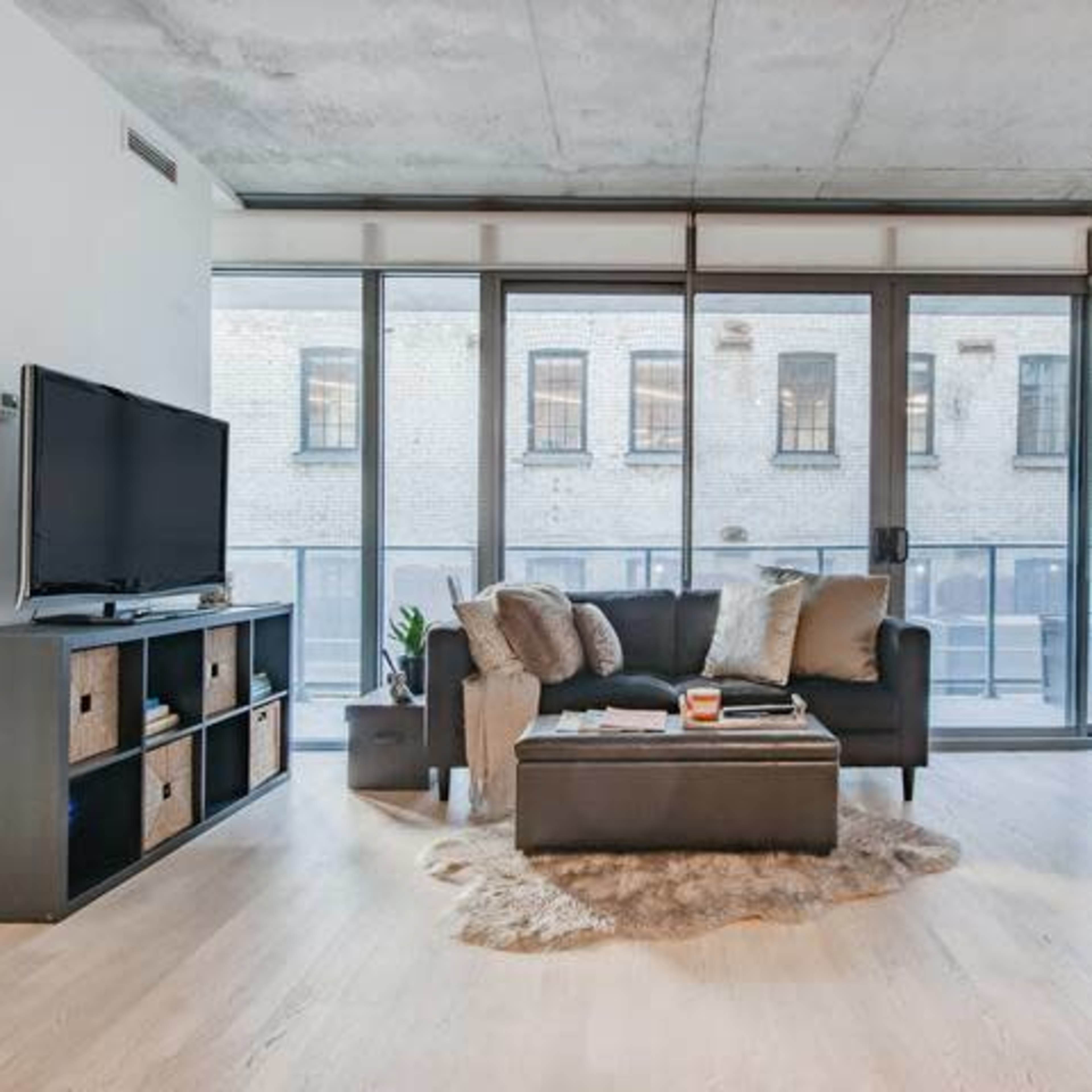 A modern living room featuring a dark couch, a coffee table, and a television, with large windows revealing a brick building outside.