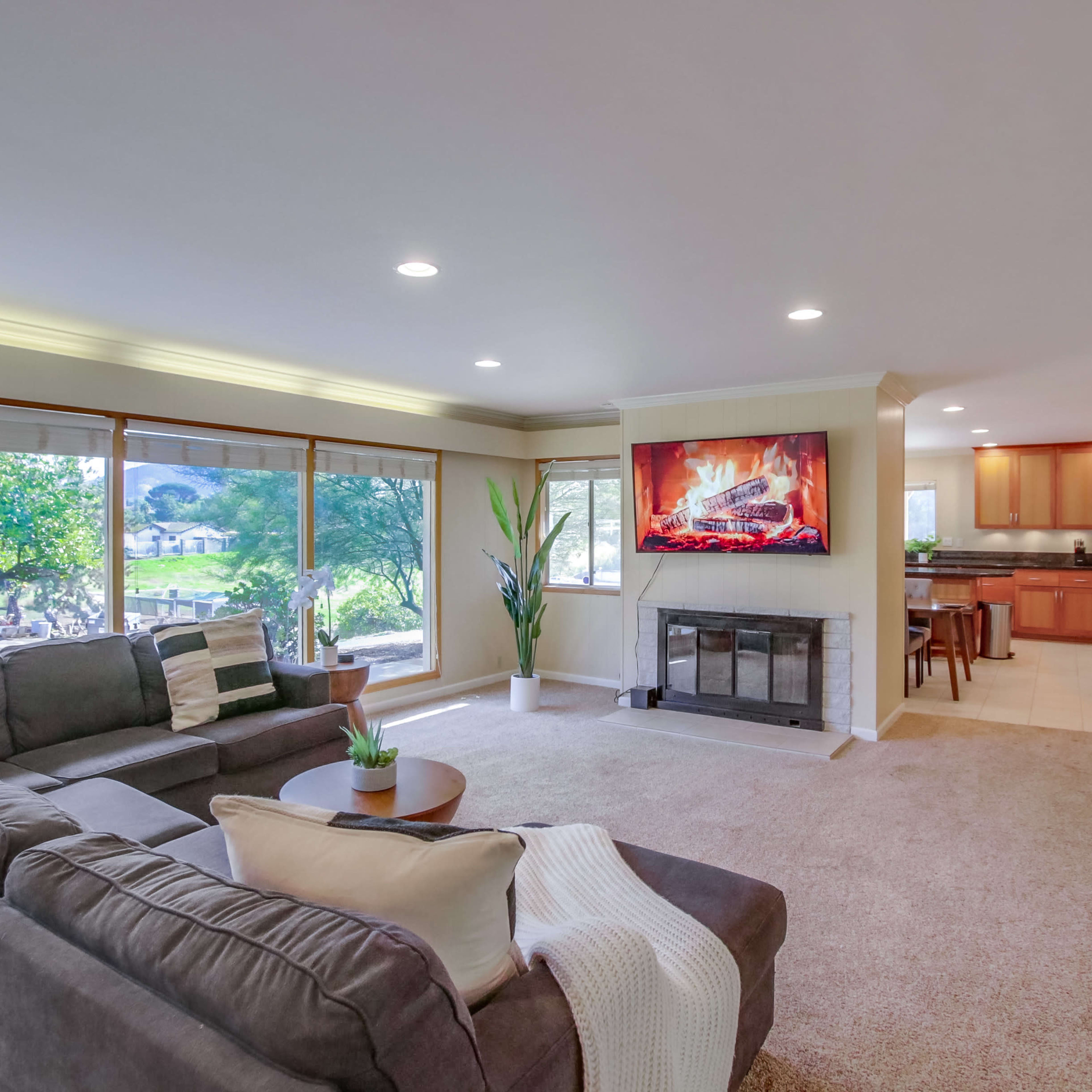 A contemporary living room with large windows, a fireplace, and an adjoining kitchen.