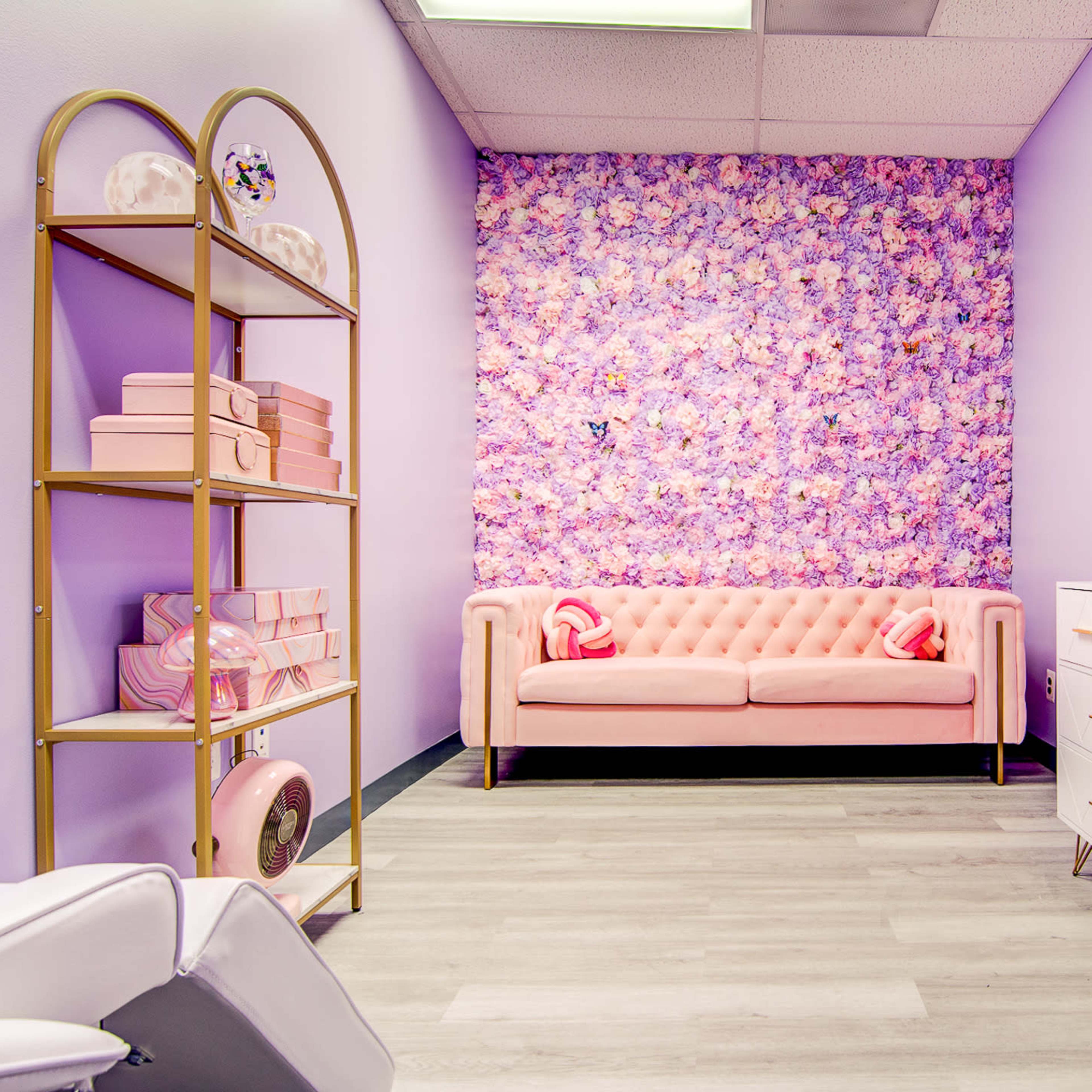 A pastel-themed room with a pink tufted sofa, a floral accent wall, a gold bookshelf, and a white dresser against one wall.