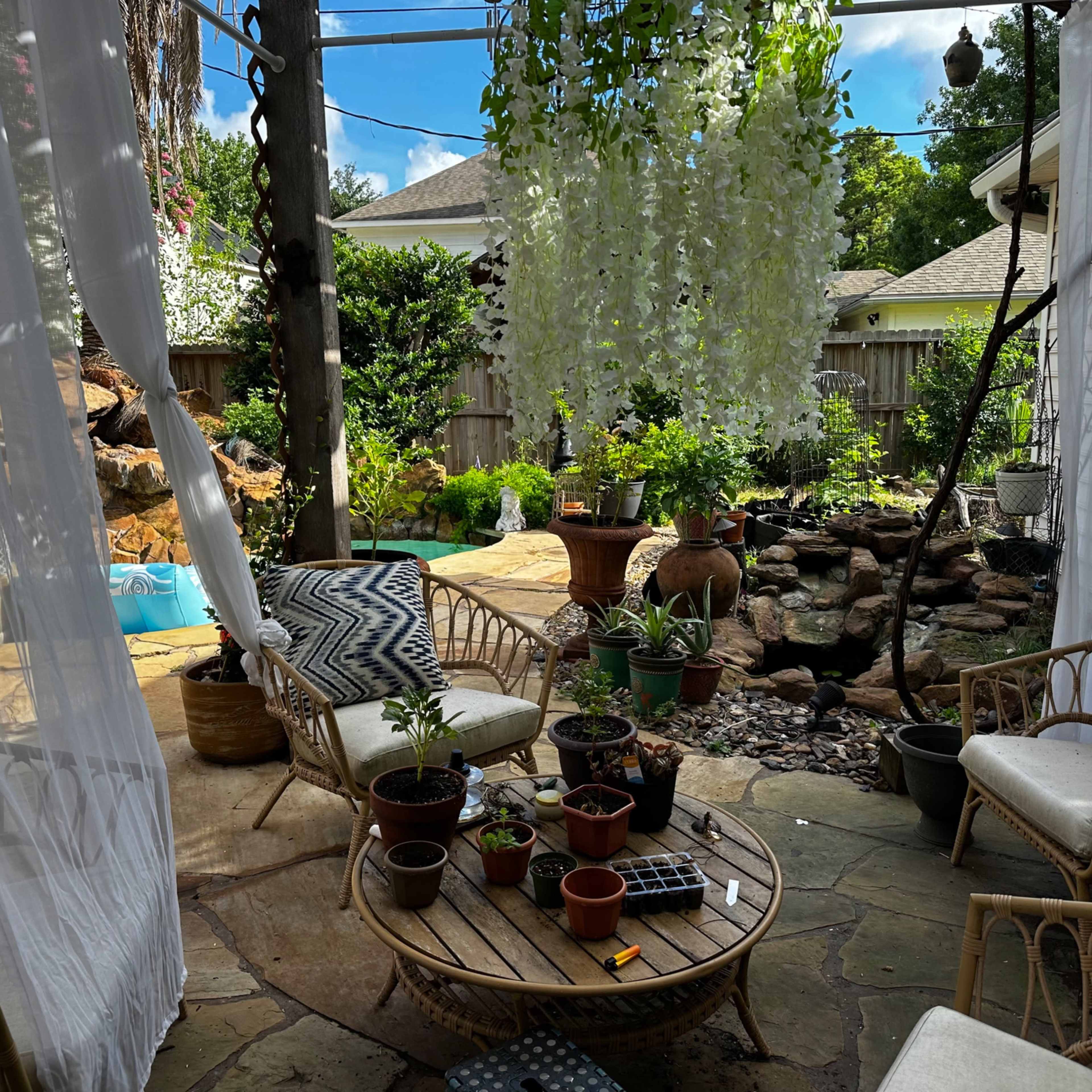 A cozy outdoor seating area features wicker chairs and a round table surrounded by potted plants and a garden backdrop.