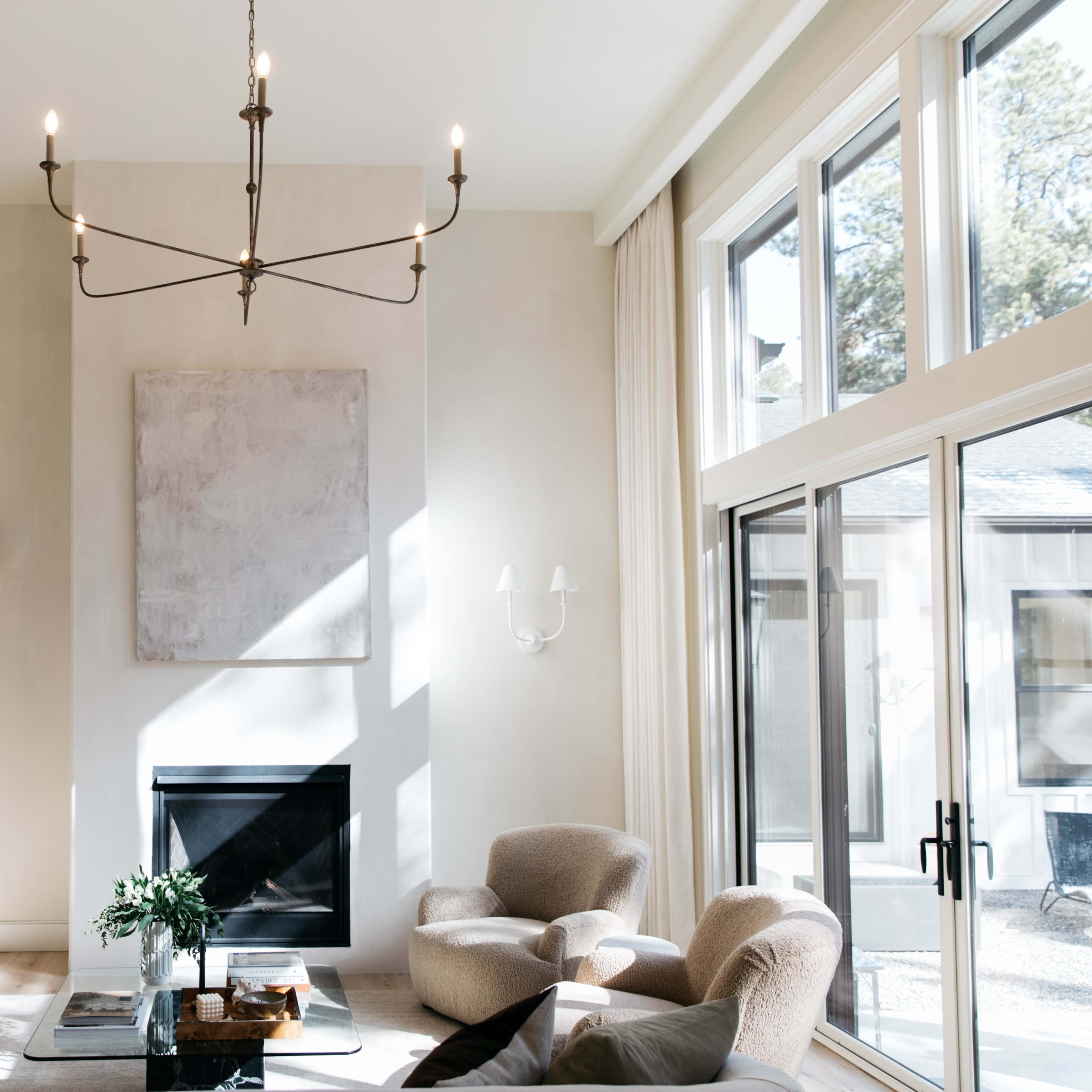 A spacious living room features a modern chandelier, a central fireplace, and large windows allowing natural light to fill the space.