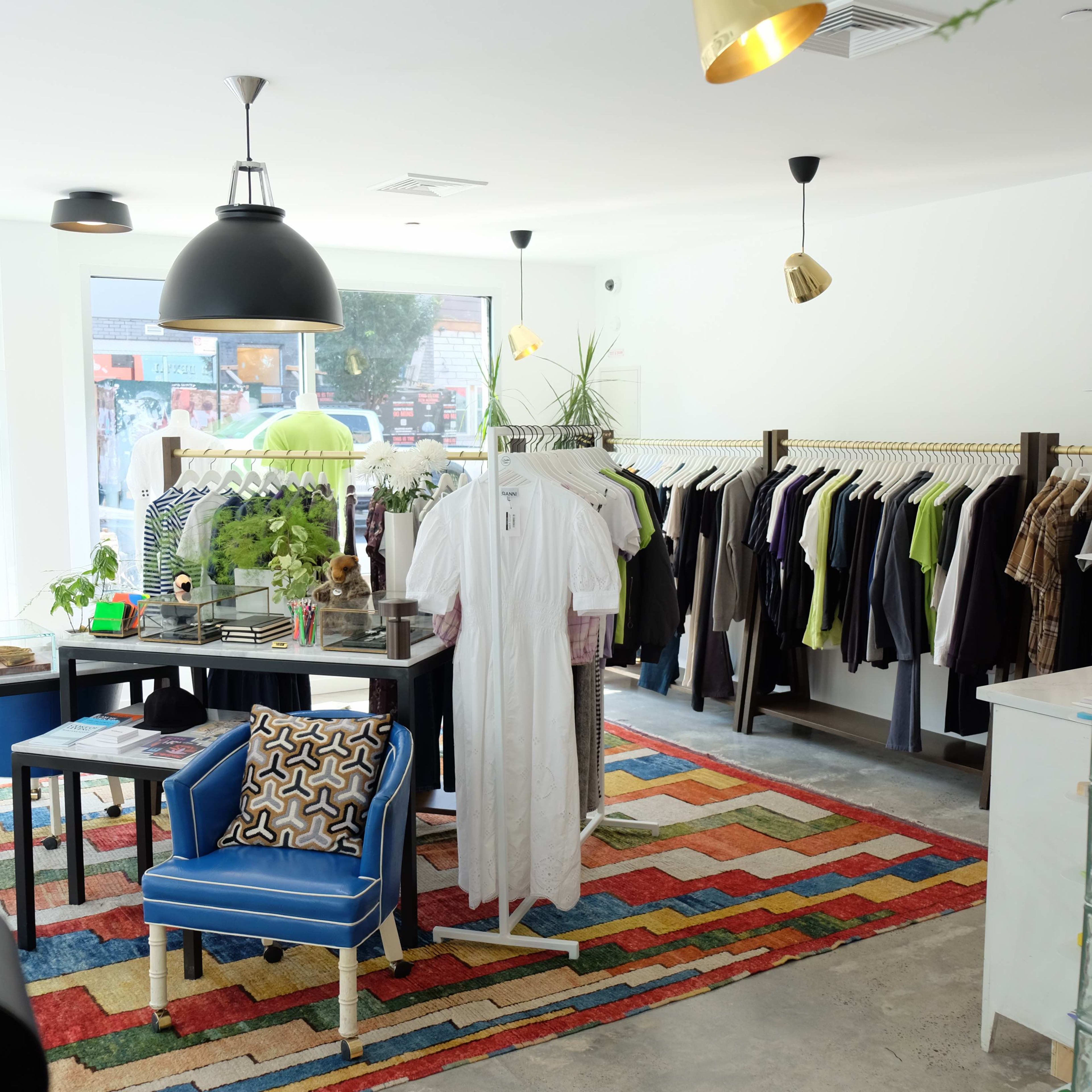 A retail clothing store features racks of apparel, a central table with decorative items, and a blue chair on a patterned rug.