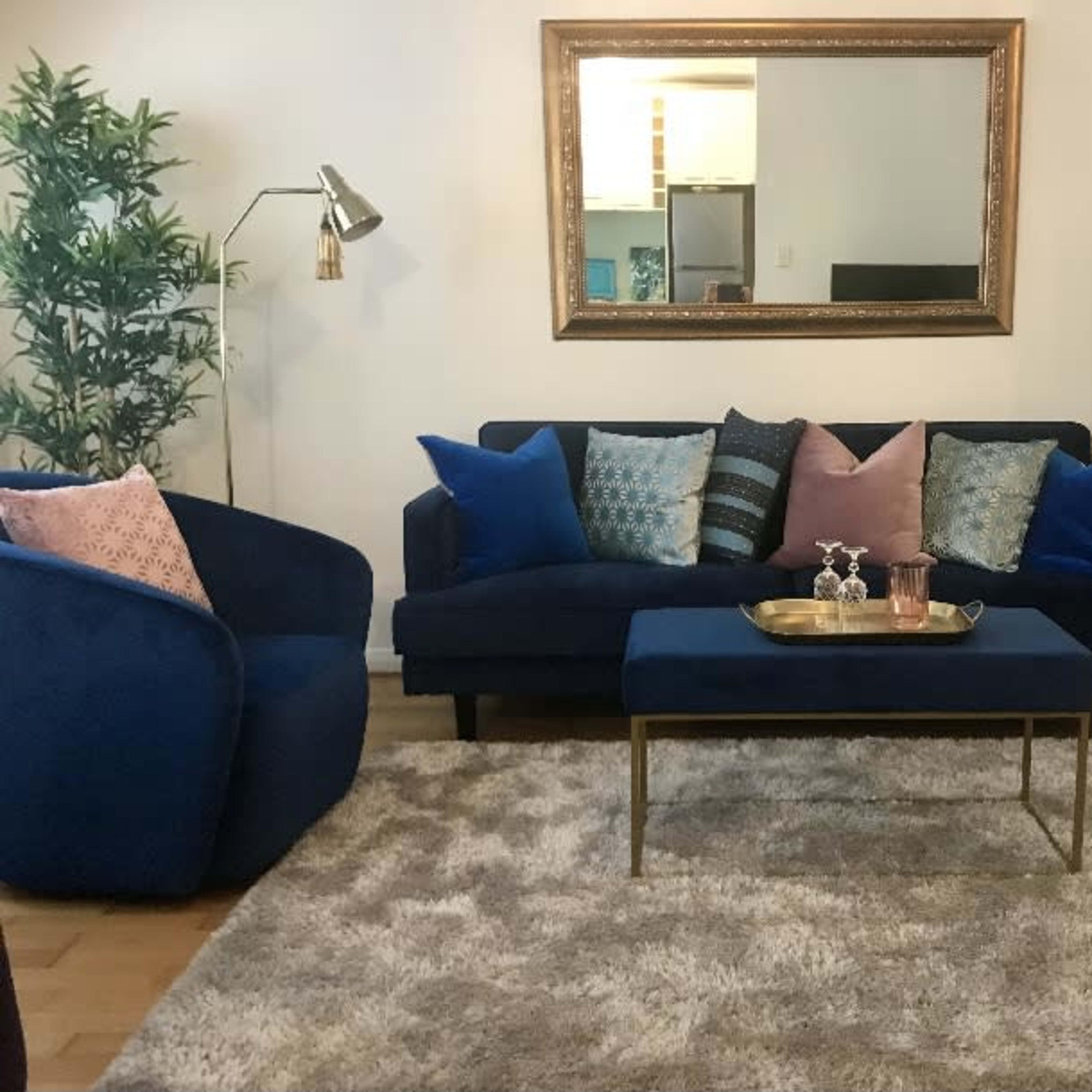 A living room features a navy blue sofa with decorative pillows, a small coffee table with a tray, and a large mirror above it, all arranged on a soft rug.