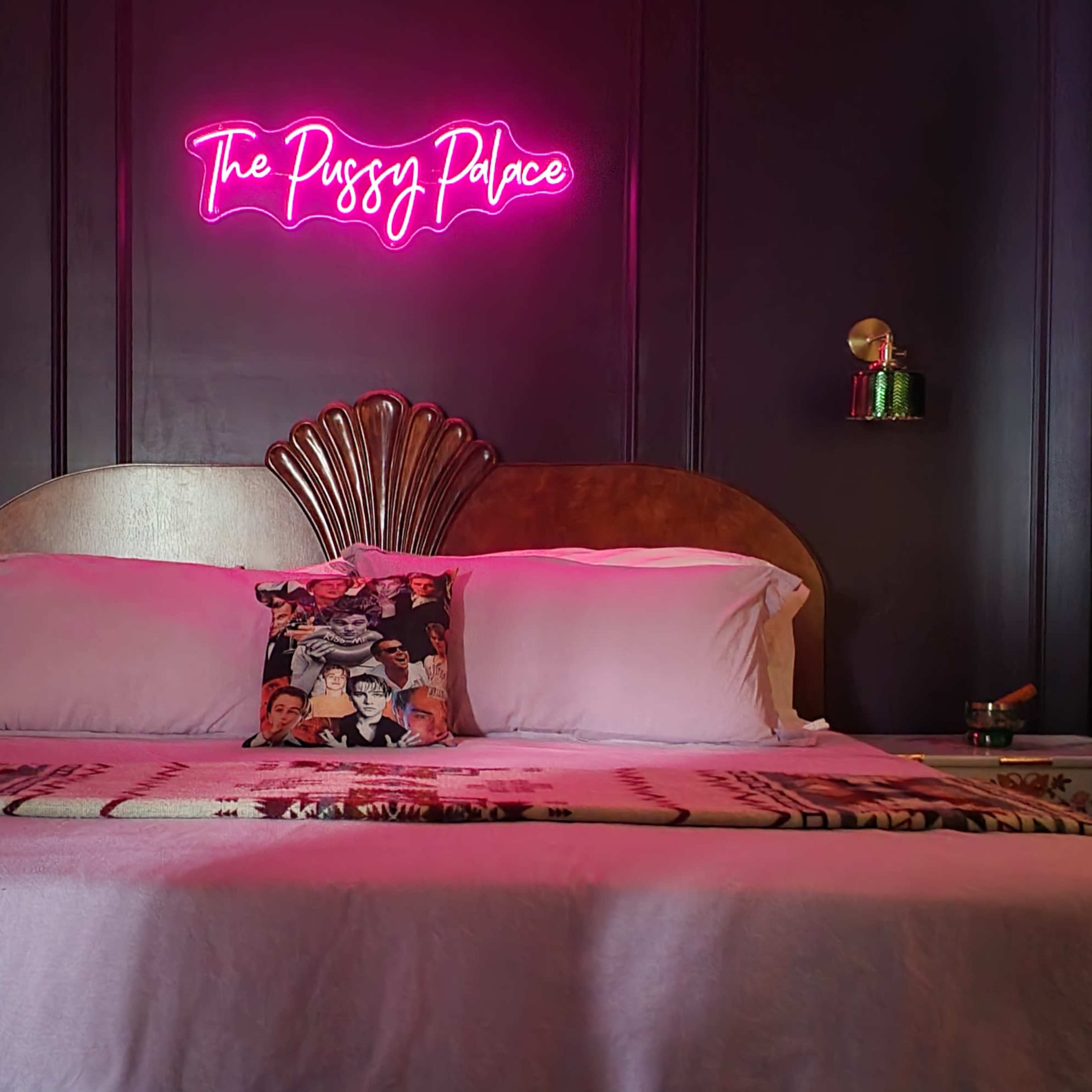 A bed with a decorative headboard is centered against a dark wall, featuring a neon sign reading "The Pussy Palace" above it.