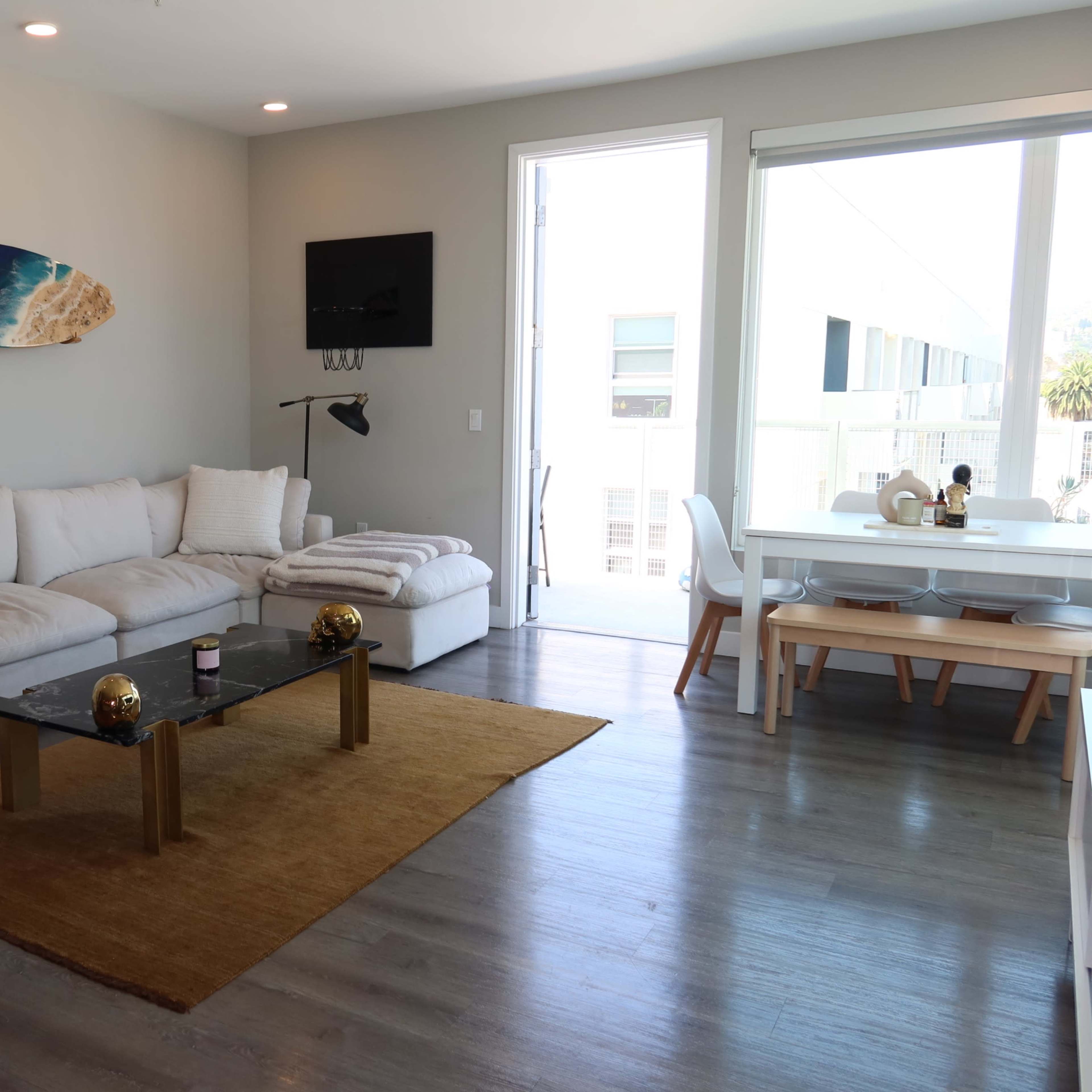 A modern living room with a white couch, a glass coffee table, a rug, and a dining area featuring a circular table and chairs next to a large window.