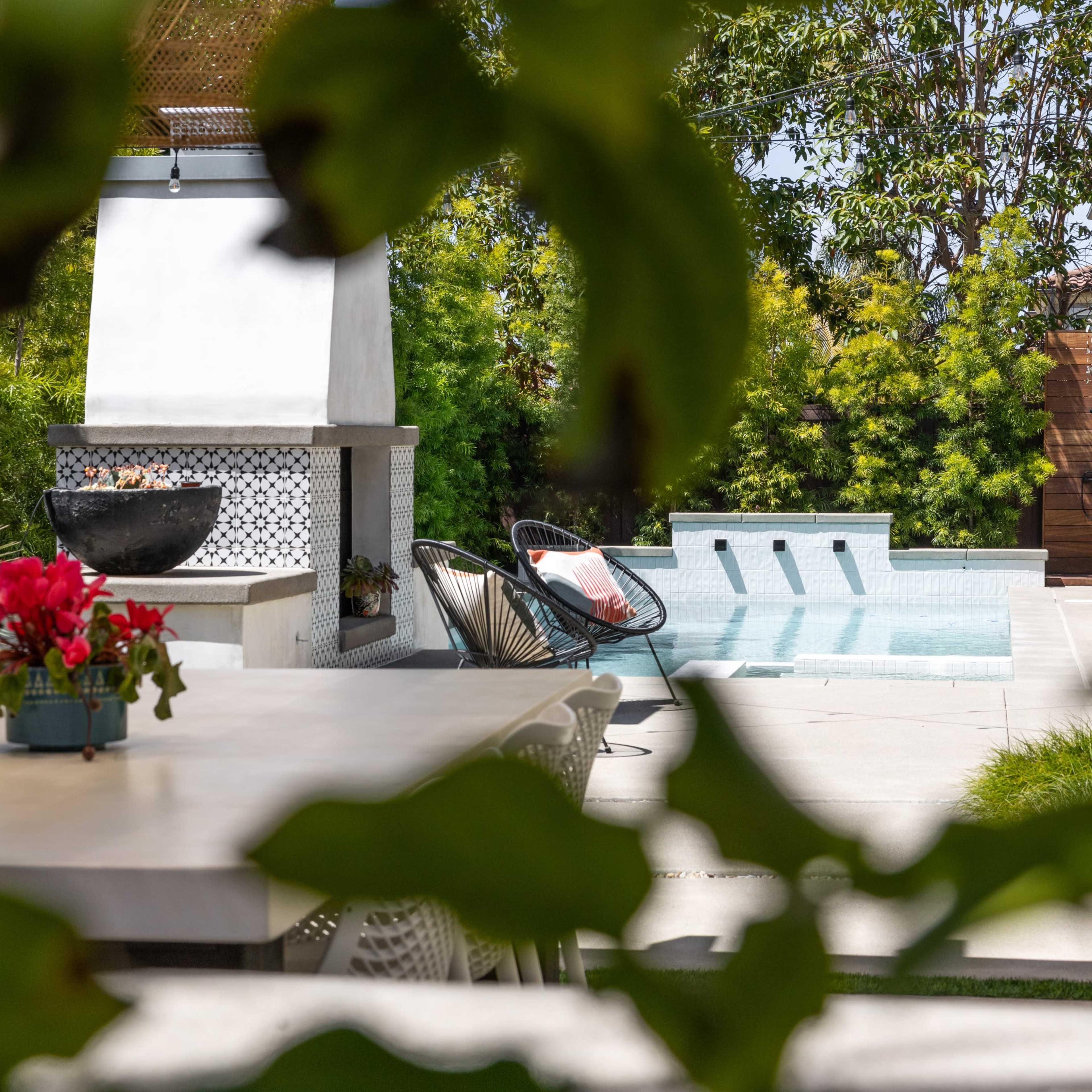 A patio features a dining table and a pool surrounded by greenery and a barbecue area.