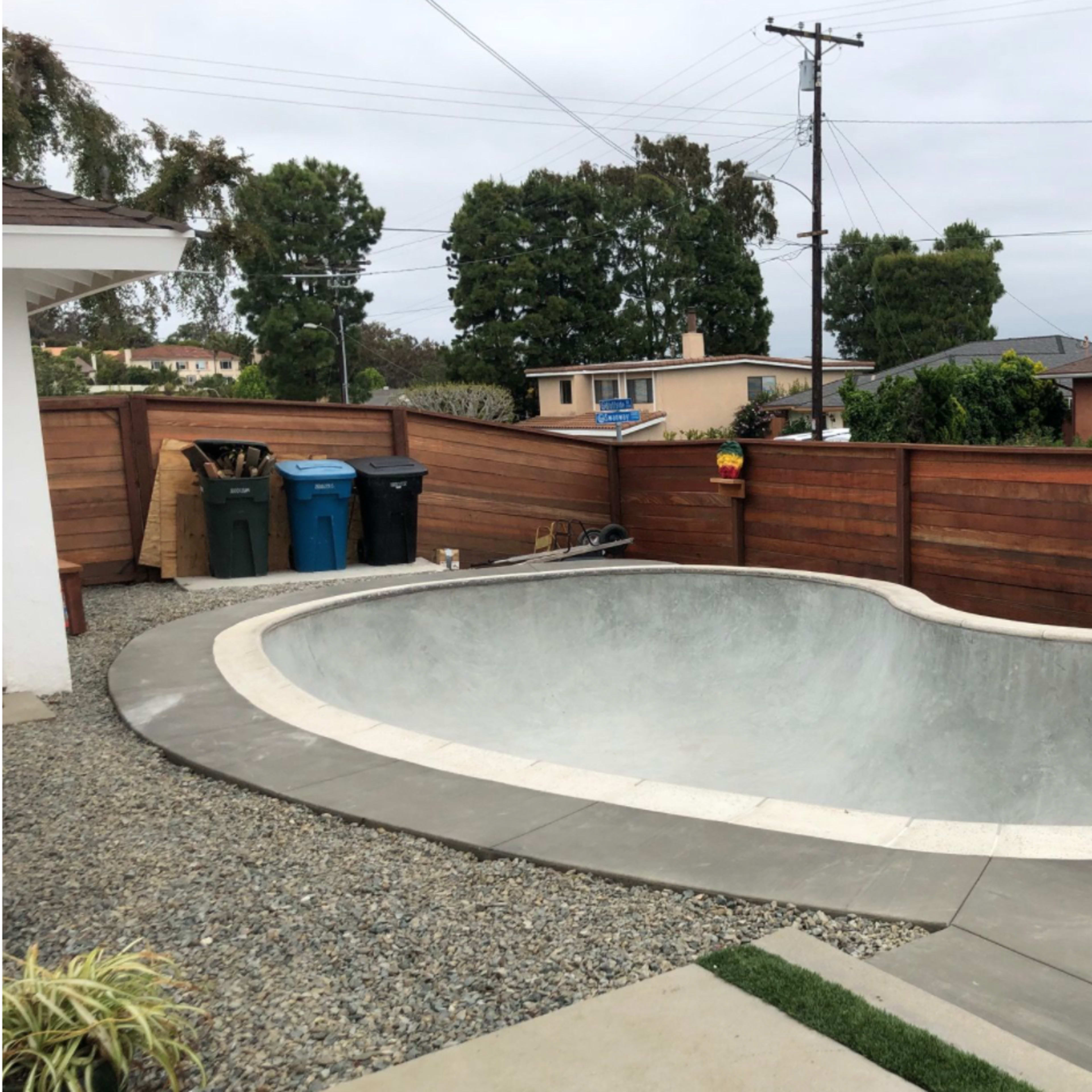 An empty, unfinished concrete swimming pool surrounded by a fenced yard with a gravel pathway and garbage bins nearby.