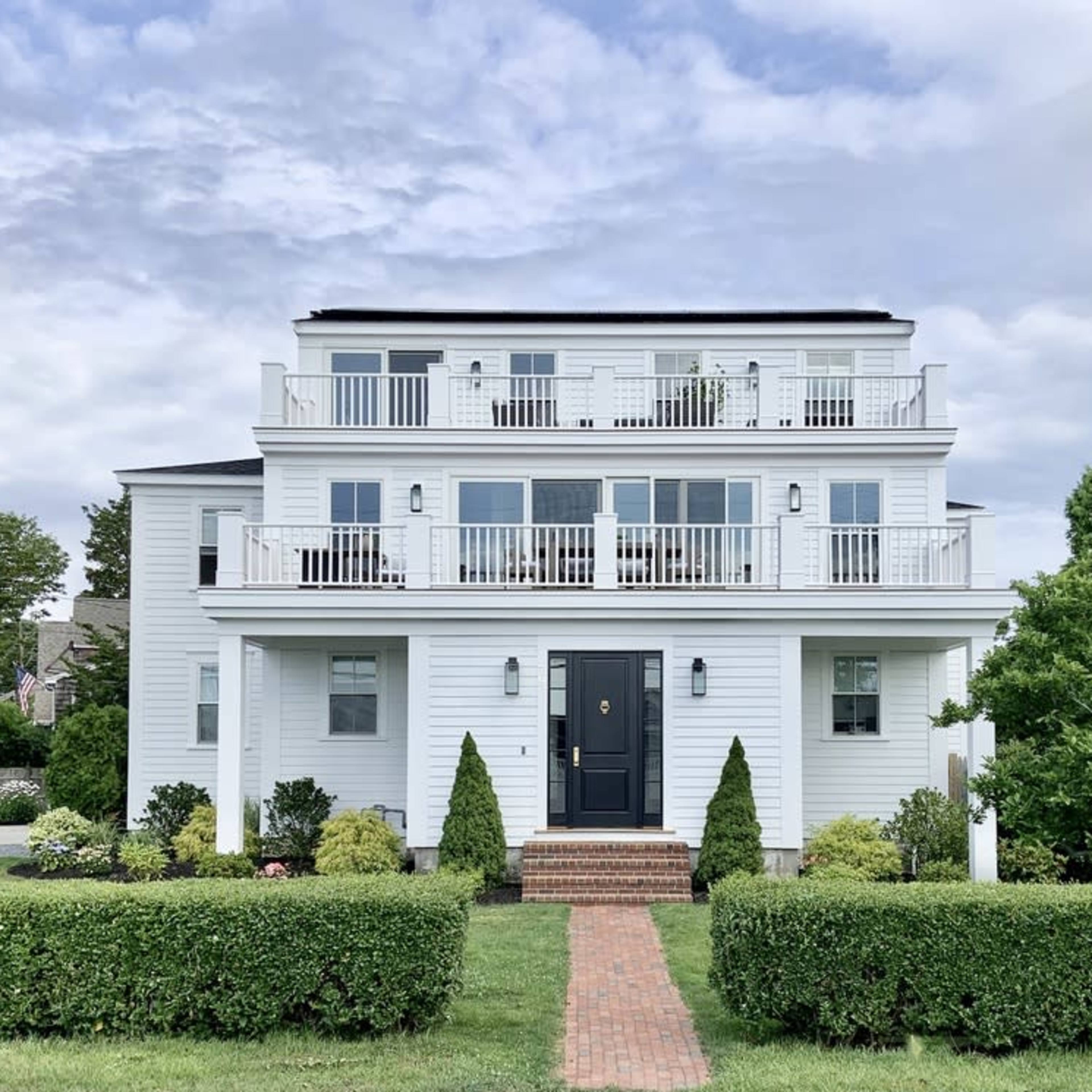 A three-story white house with balconies on each floor is flanked by neatly trimmed hedges and a brick pathway leading to the front door.