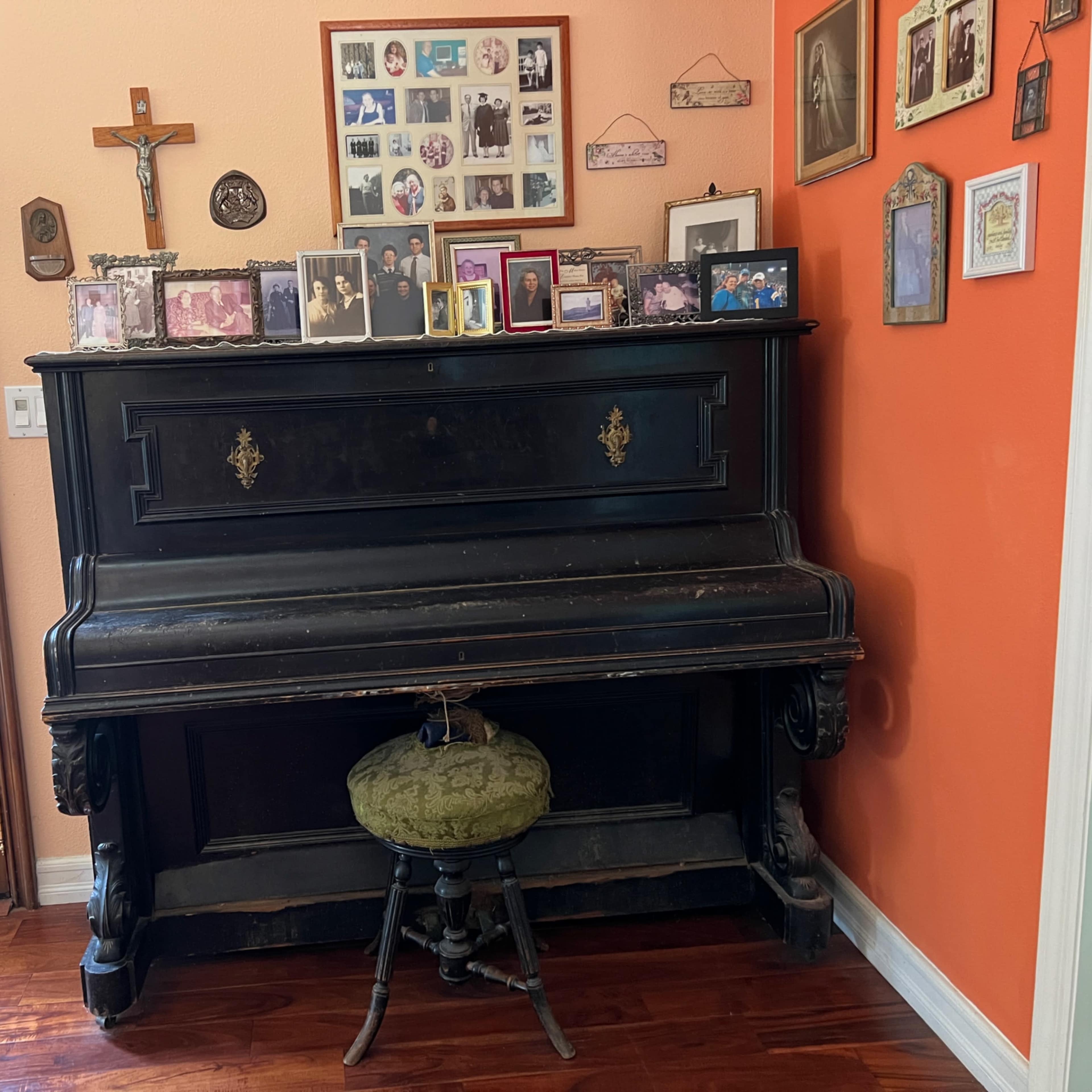 An antique piano is positioned against a wall adorned with a variety of framed photographs and a crucifix.