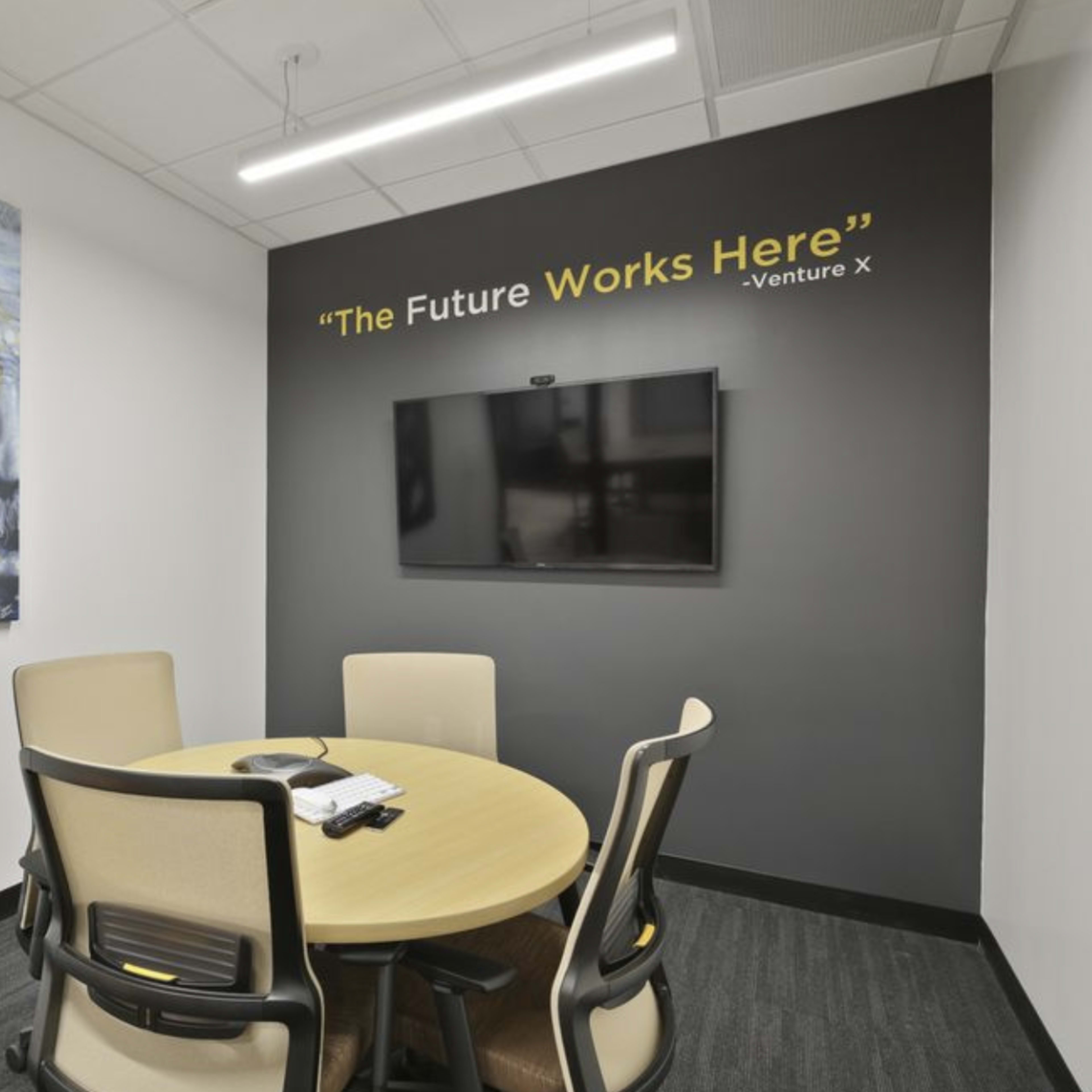 A small conference room features a round table with four chairs, a wall-mounted television, and a portrait of Albert Einstein, accompanied by the phrase "The Future Works Here" painted on the wall.