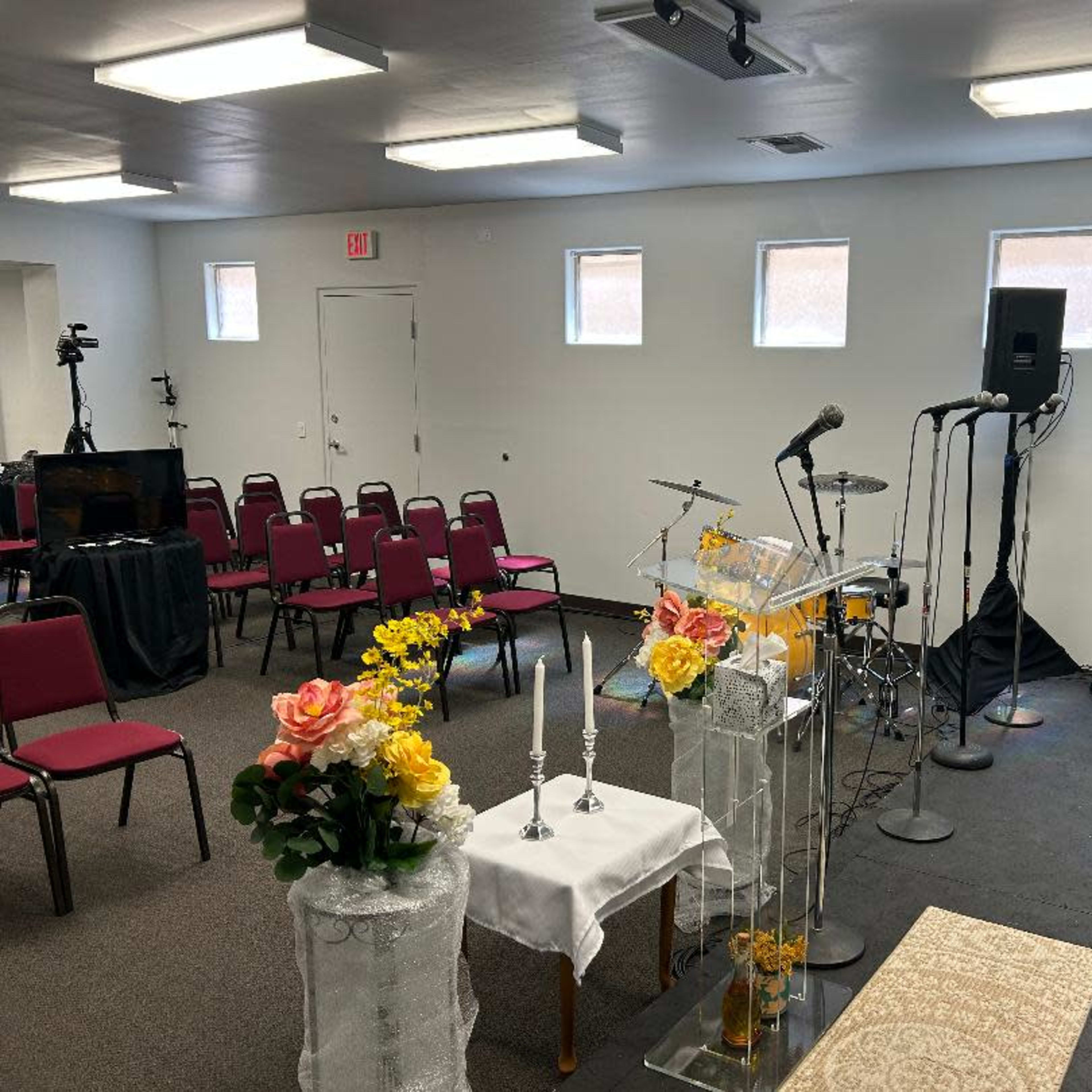 A small room arranged with chairs for an event, featuring a stage setup with microphones, musical instruments, and decorative floral arrangements.