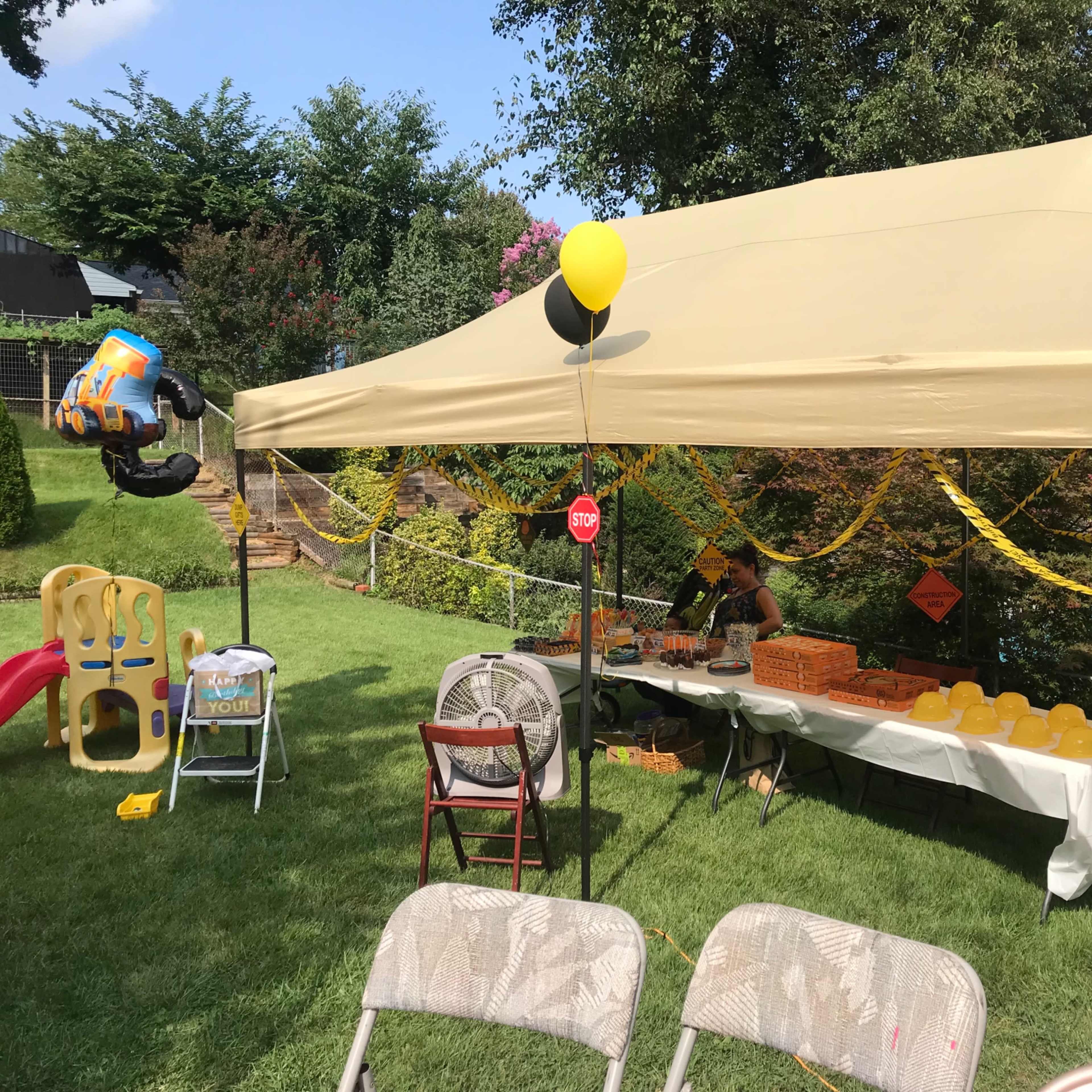 A large tent is set up in a grassy area, with a table displaying yellow balloons and treats, surrounded by chairs and decorations.