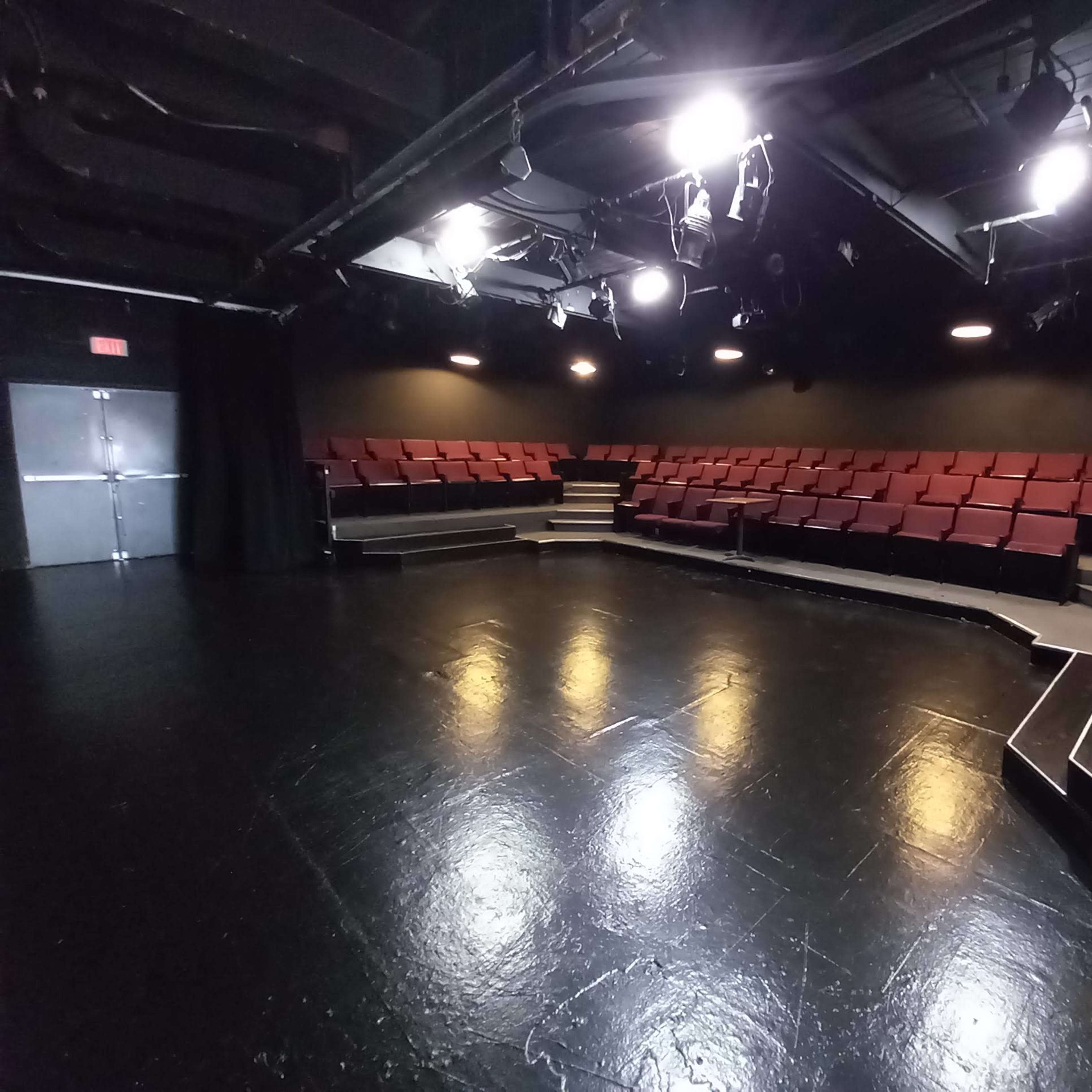 An empty theater with red seats arranged in a semi-circular layout around a dark, polished stage.
