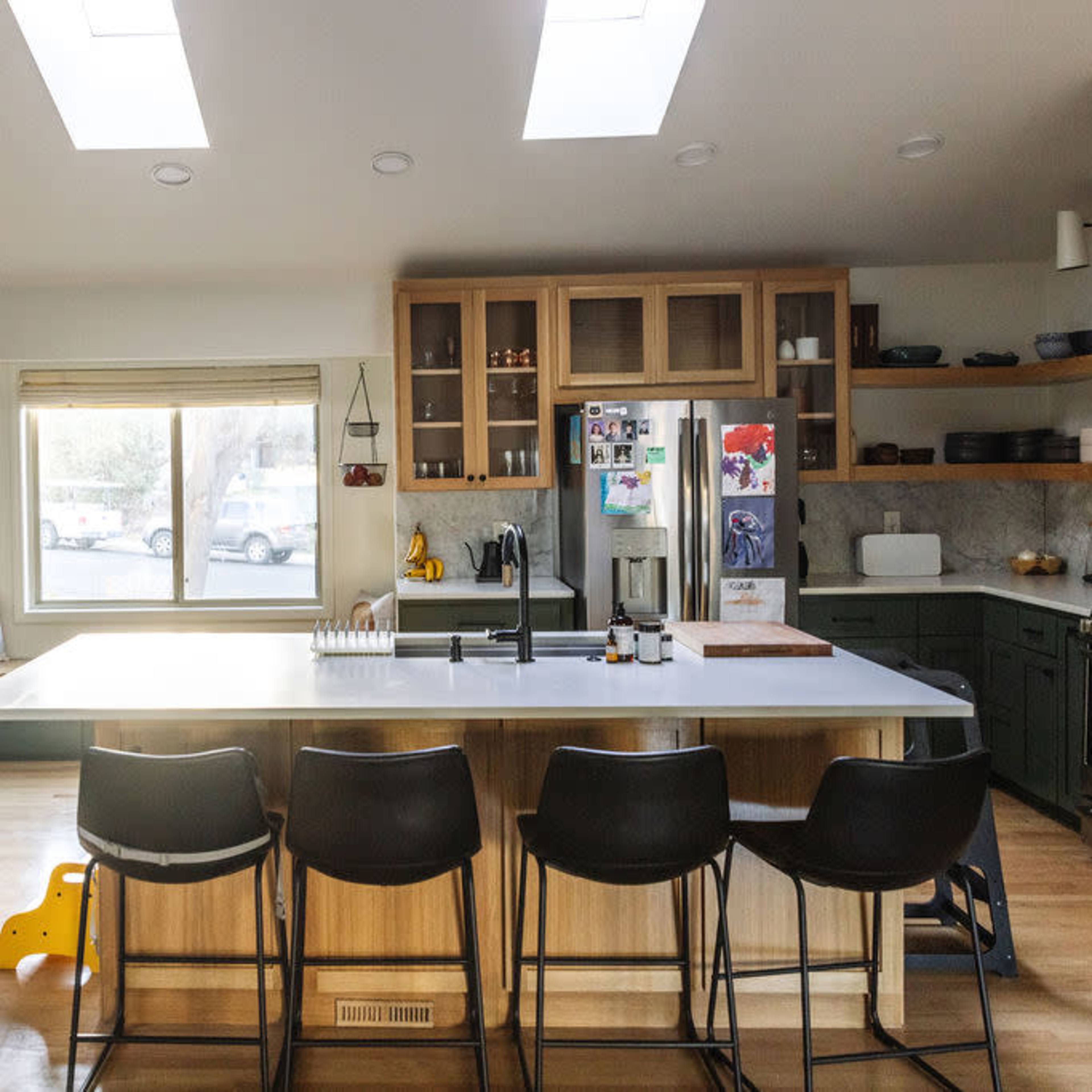 A modern kitchen features green cabinetry, a central island with bar stools, and multiple skylights allowing natural light to enter the space.