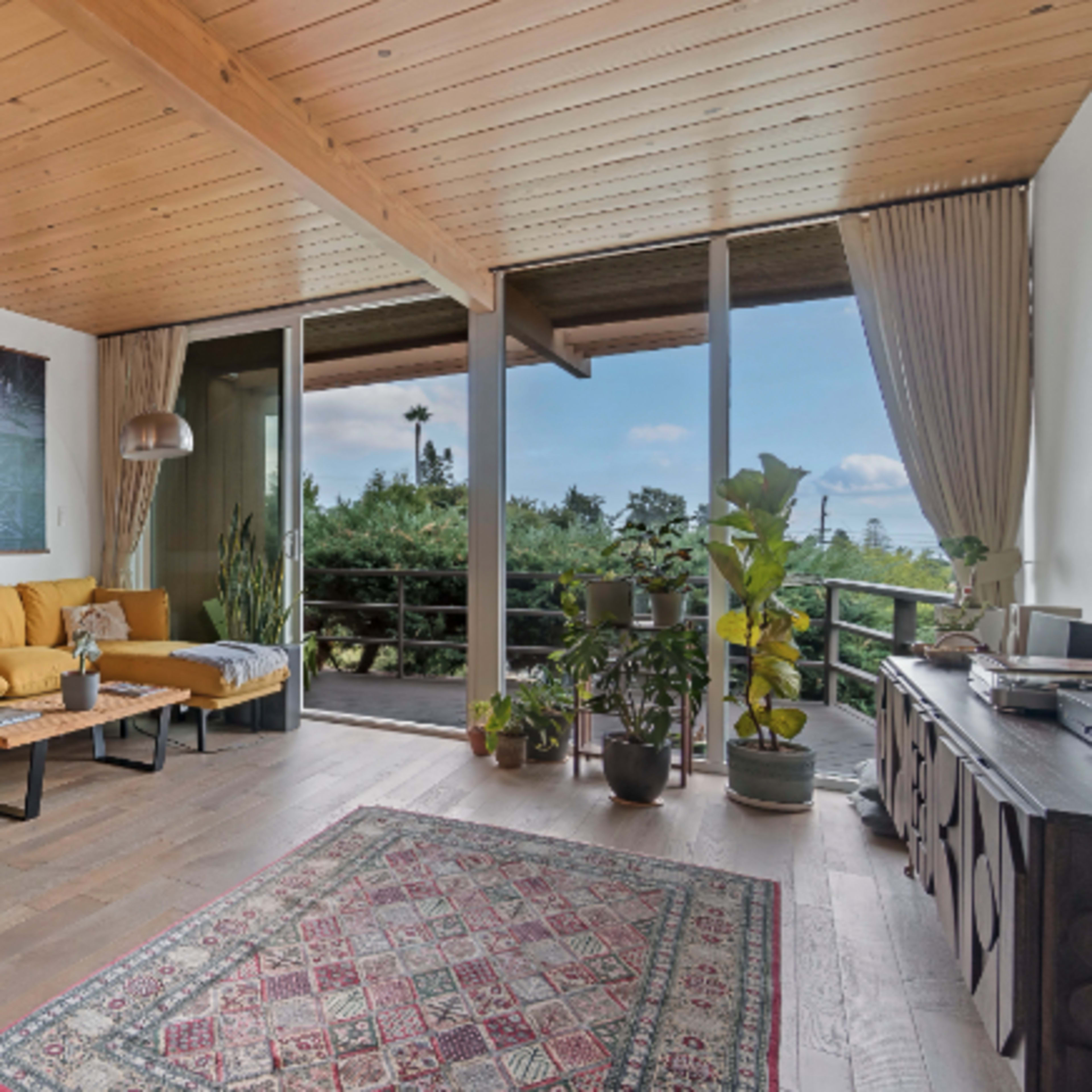A modern living space featuring a dining table near a sofa, large windows with views of greenery, and various plants around the room.