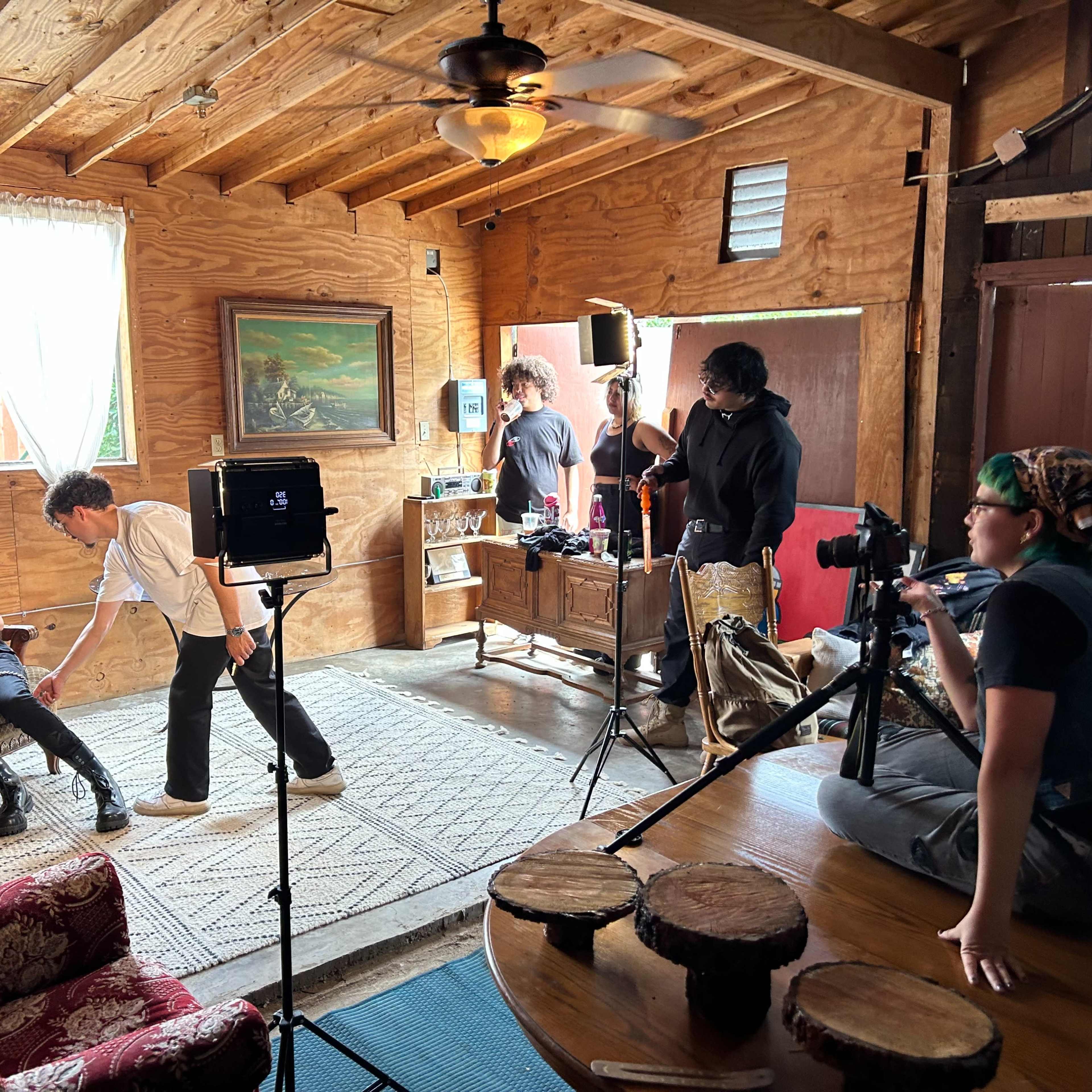 A film crew is set up in a wooden cabin interior, with one person being filmed while others adjust equipment and prepare for a shoot.