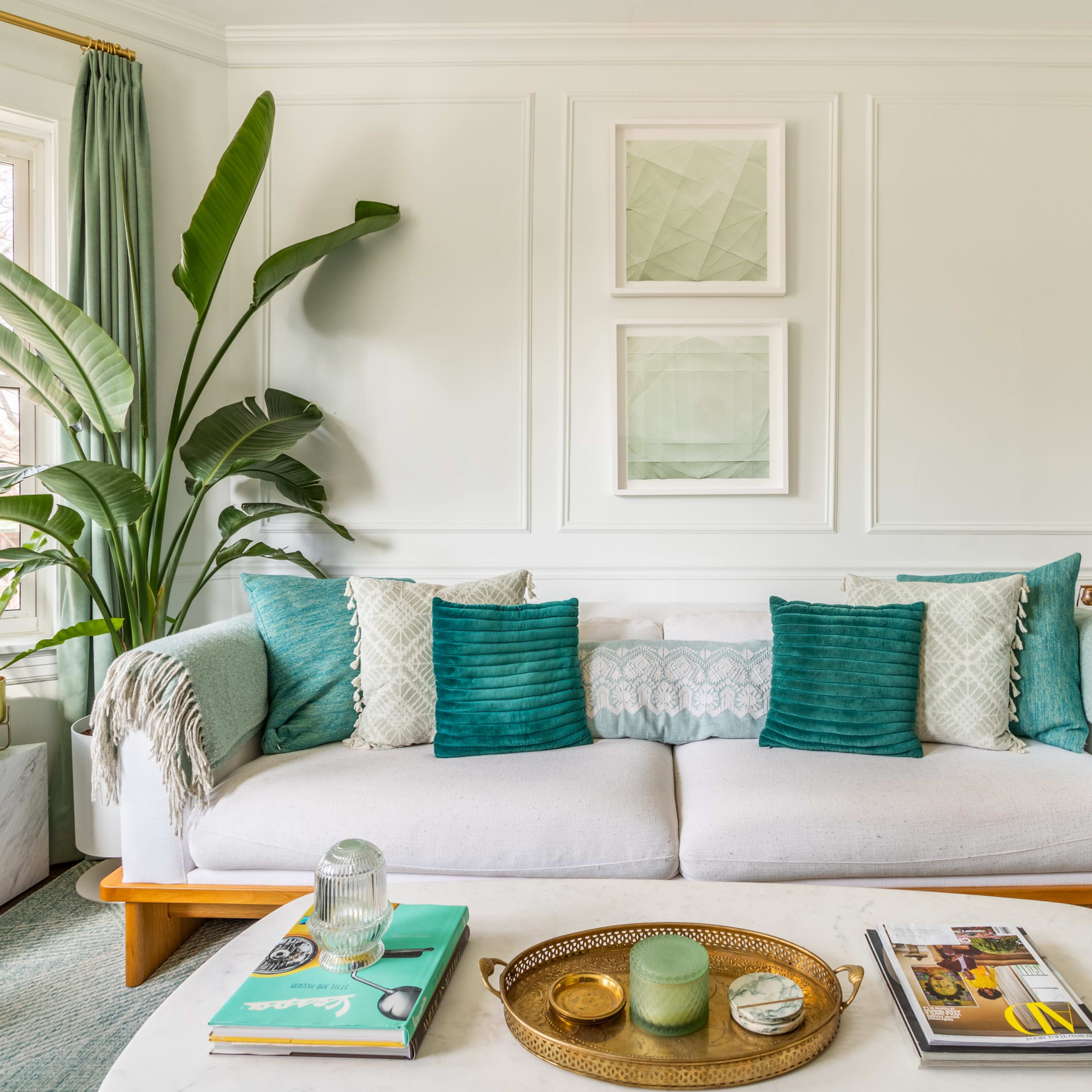 A bright living room features a white sofa adorned with teal pillows, surrounded by green plants and decorative art on the walls.