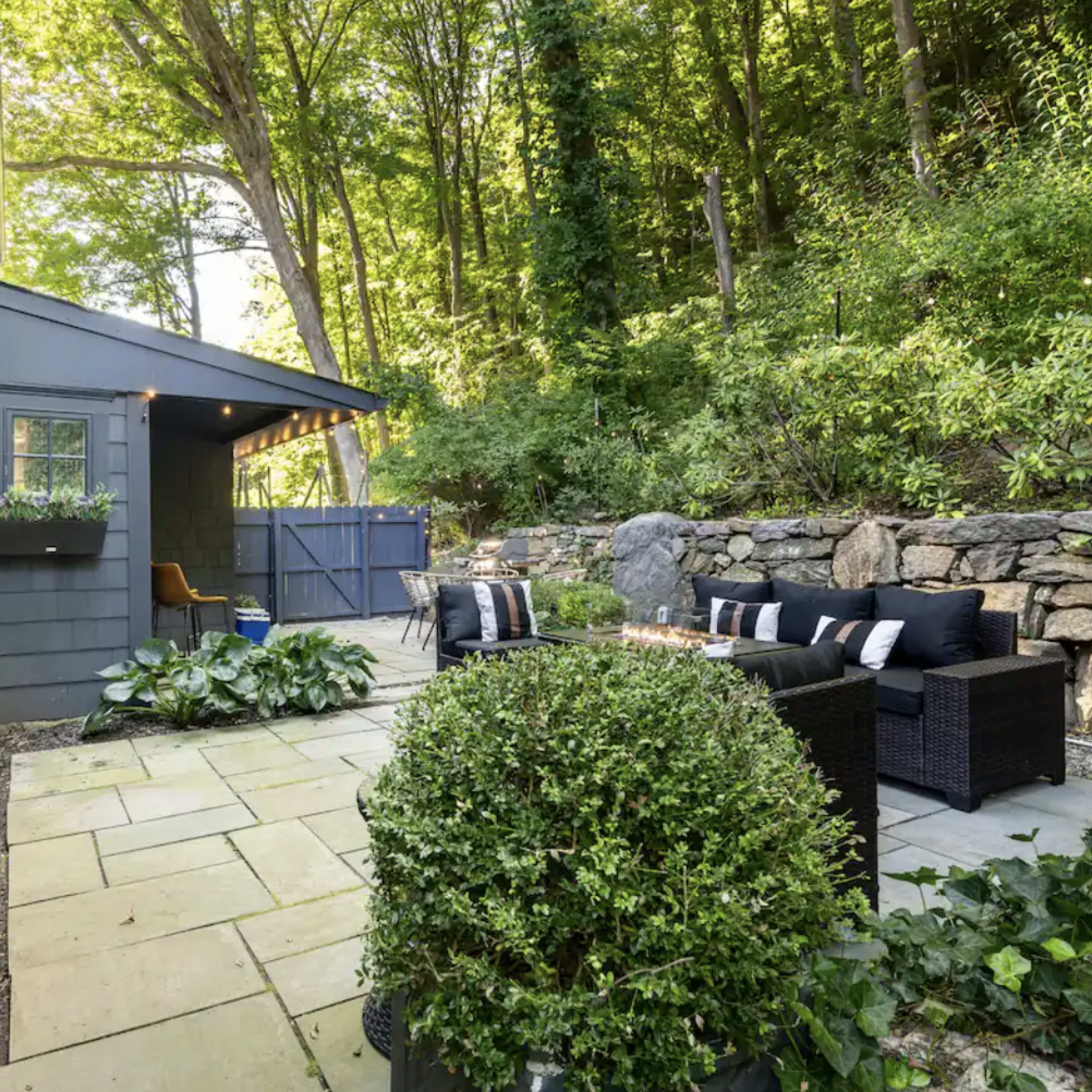 A stone patio features outdoor seating surrounded by lush greenery and a pathway leading to a wooden gate.