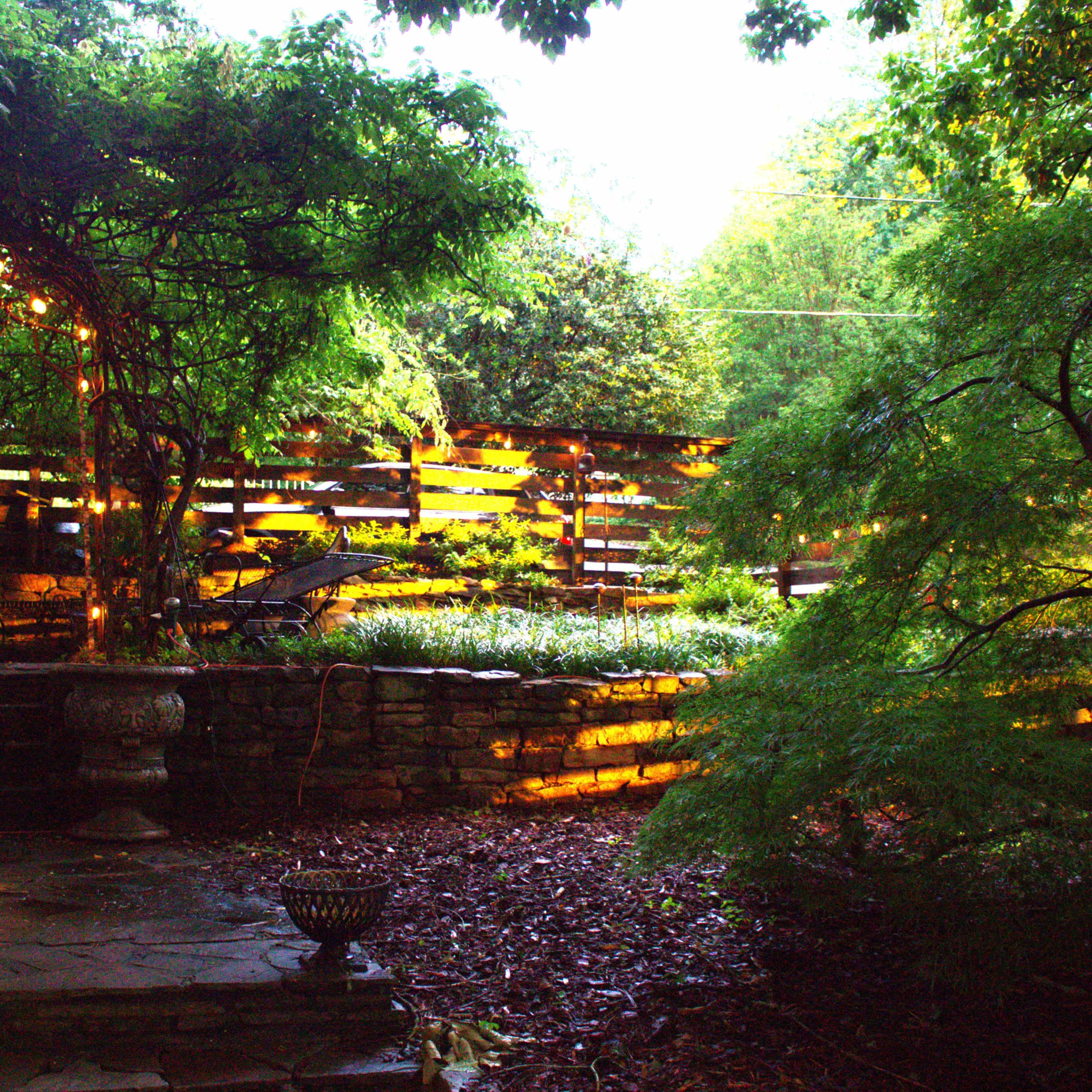 A stone pathway leads to a garden area illuminated by hanging lights, surrounded by lush greenery and wooden fencing.