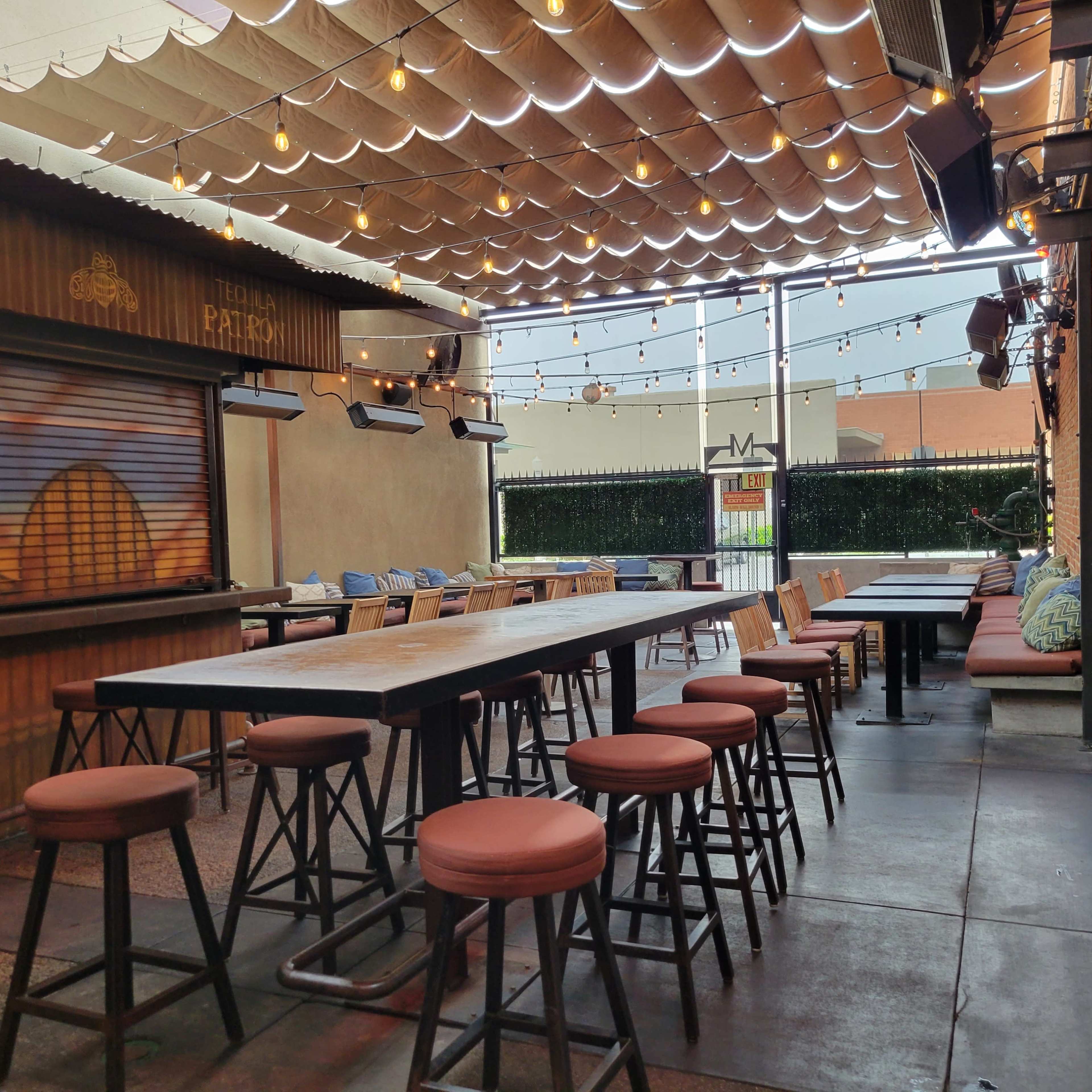An outdoor restaurant patio featuring wooden tables, high stools, and string lights overhead.