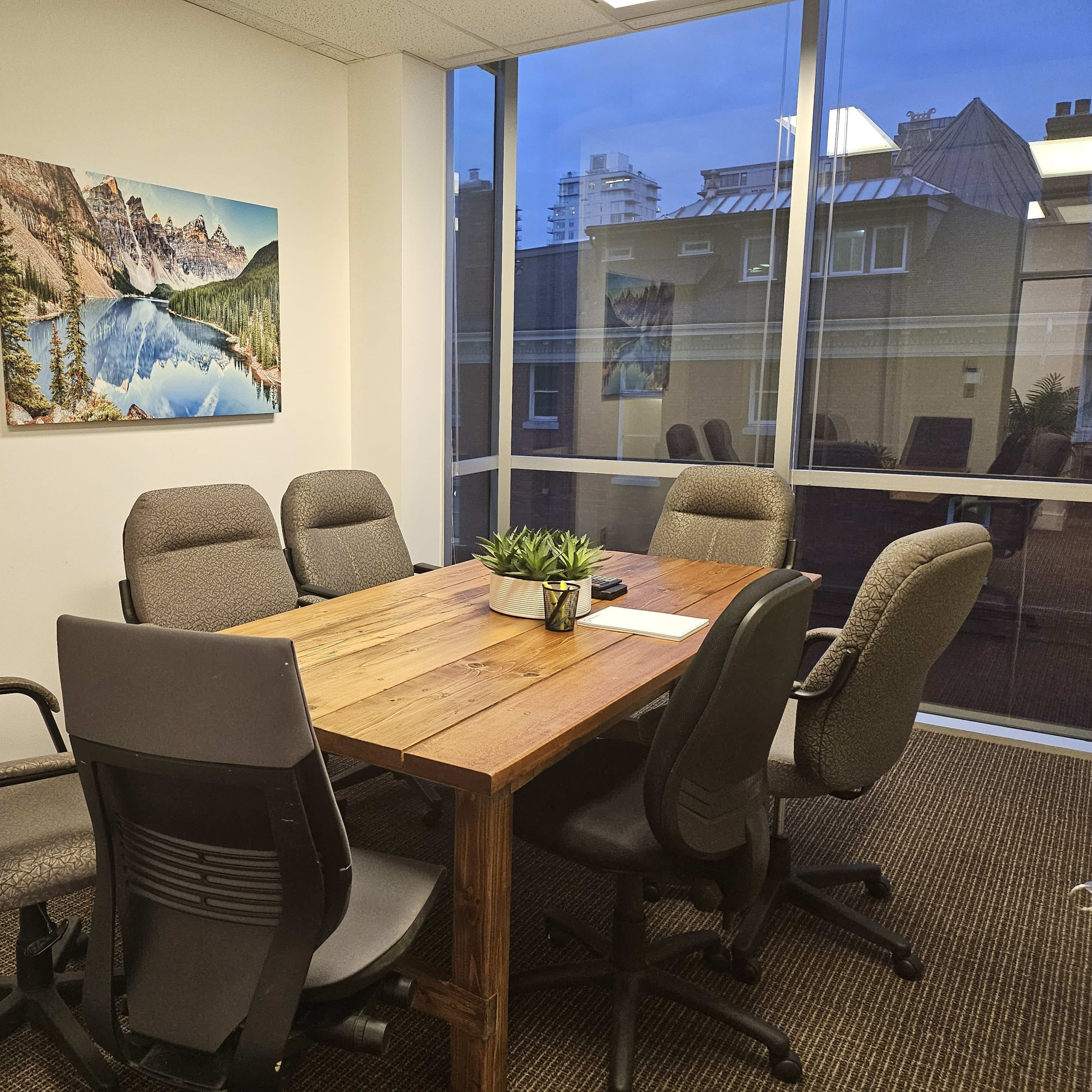 A conference room is furnished with a large wooden table surrounded by six ergonomic chairs, and a landscape artwork hangs on the wall.