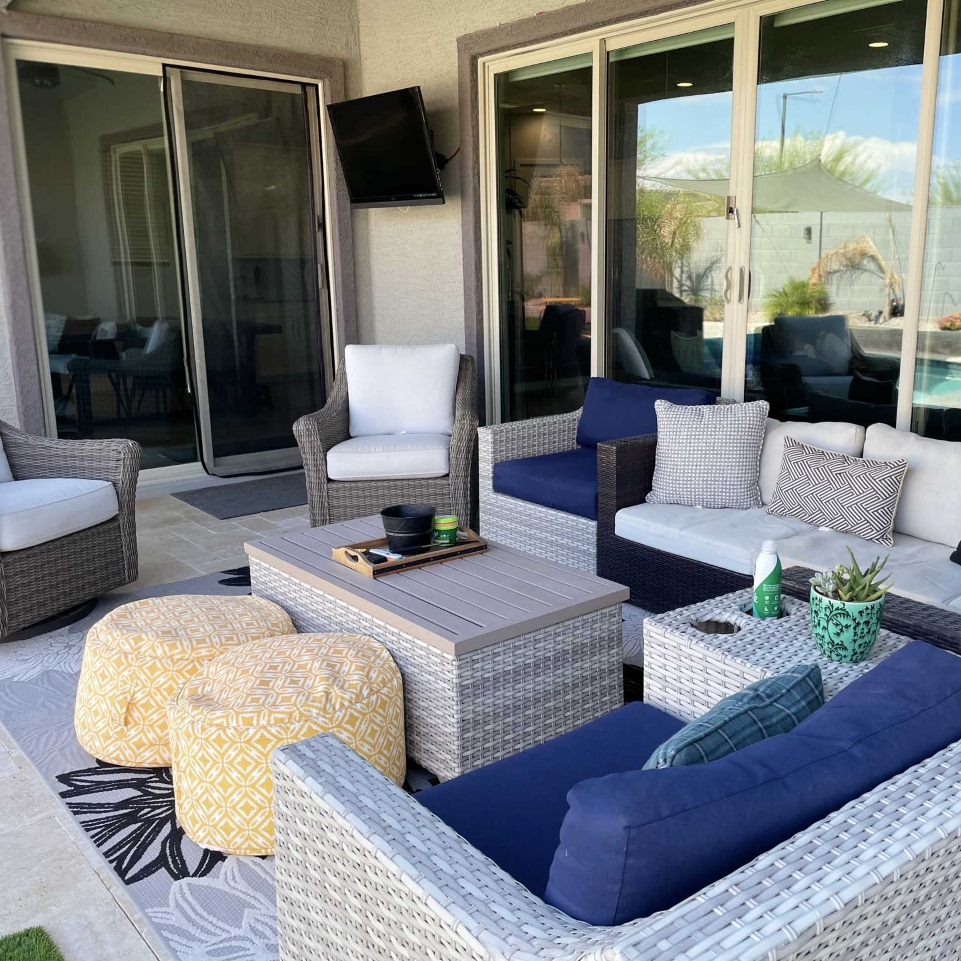 A modern outdoor seating area features wicker furniture, colorful cushions, and a central table, surrounded by large windows that overlook a pool.