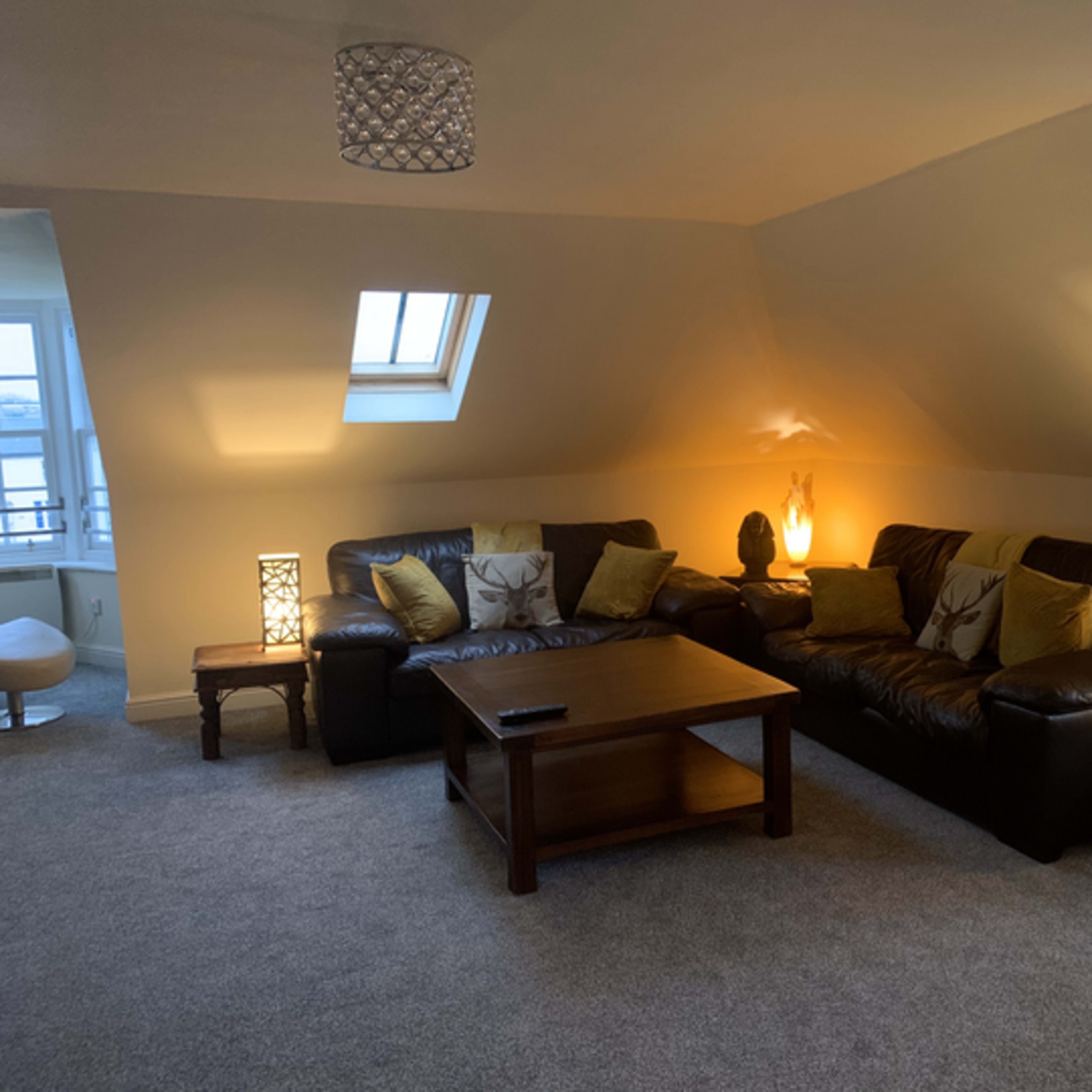A cozy living room in an attic, featuring two dark leather sofas facing a wooden coffee table and illuminated by decorative lamps.