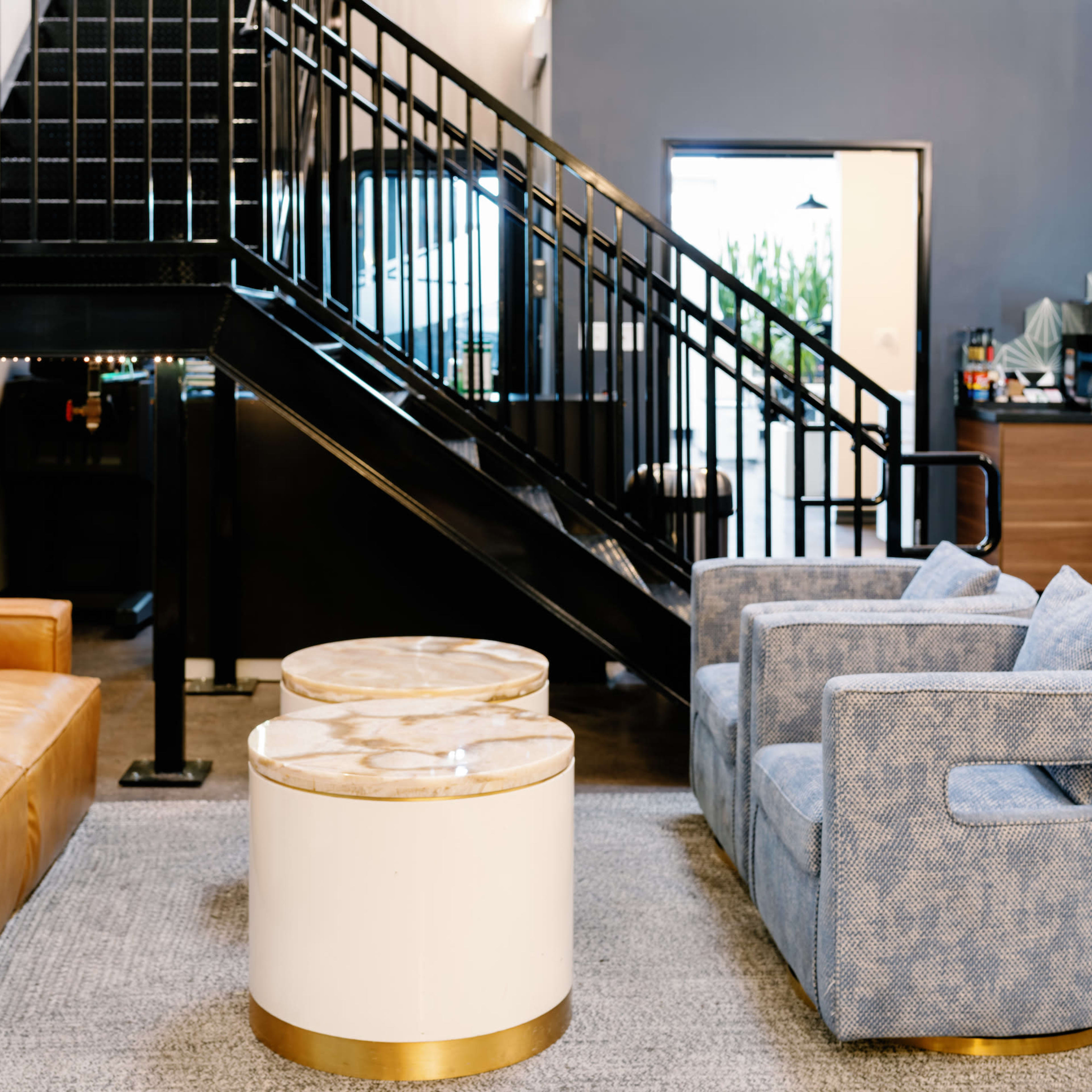 A brown leather sofa and a blue upholstered armchair are arranged across from two circular coffee tables on a gray rug under an industrial staircase.