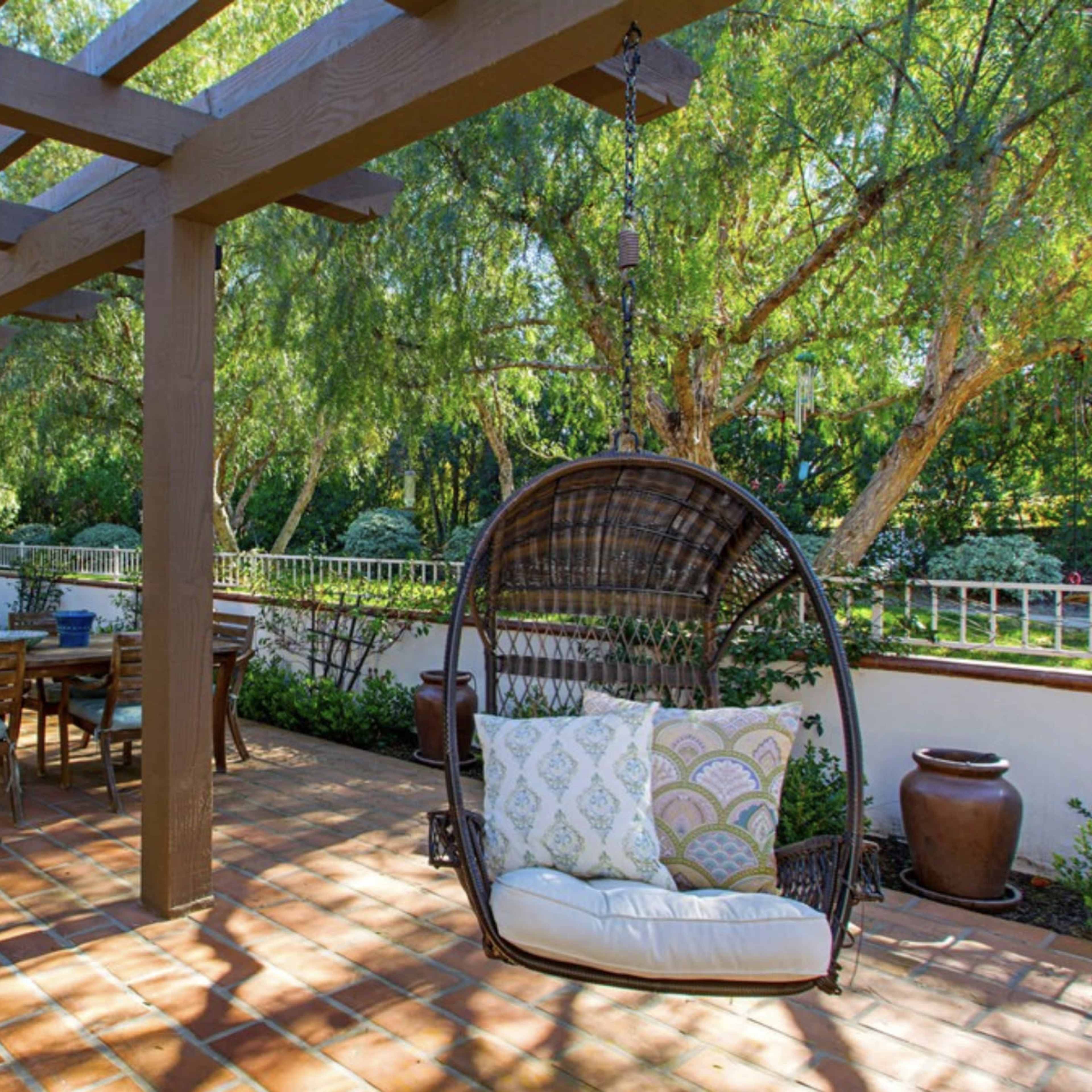 A covered patio features hanging chairs and a dining table surrounded by greenery.