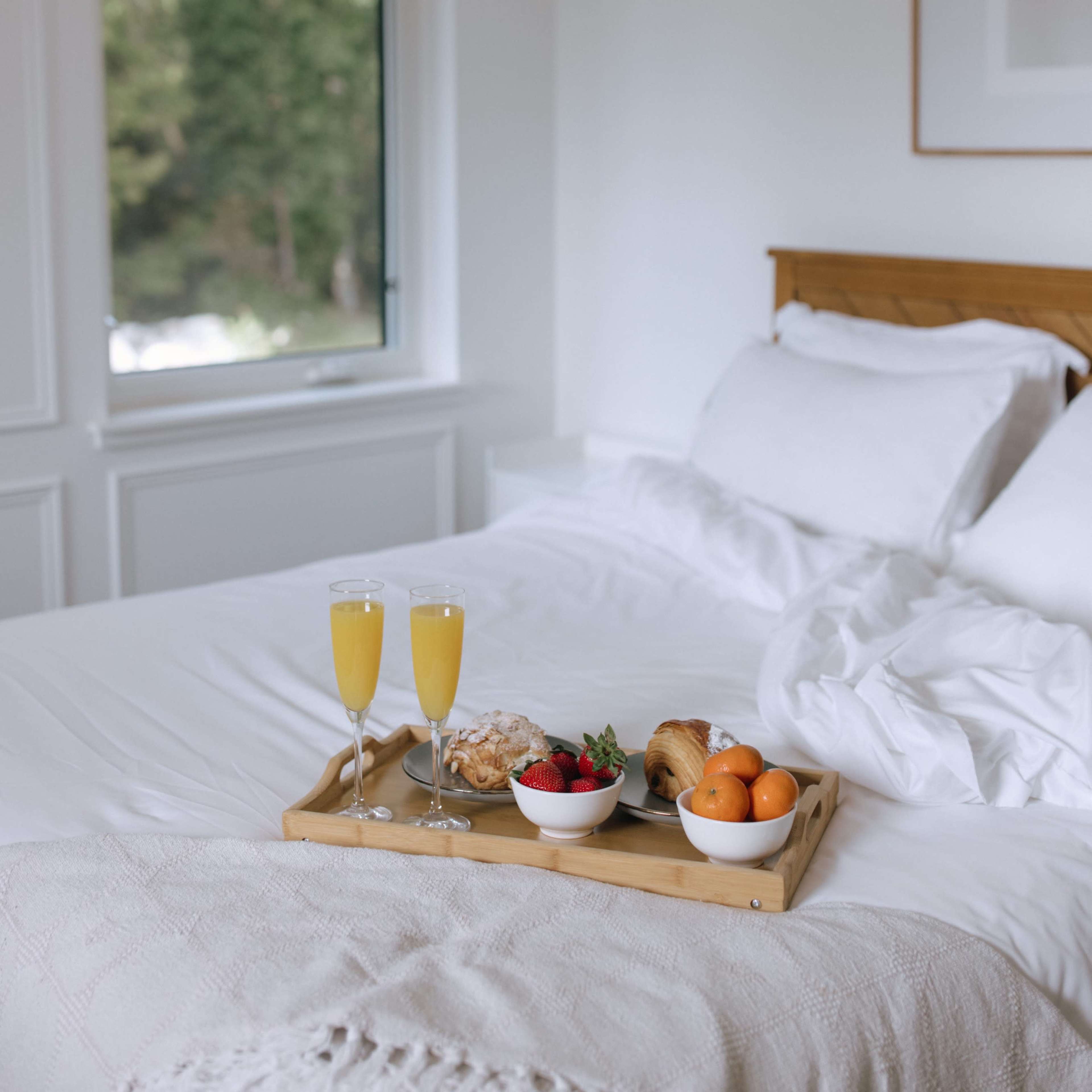 A neatly made bed with white linens is set with a wooden tray holding two glasses of orange juice and a variety of snacks, positioned near a large window overlooking greenery.