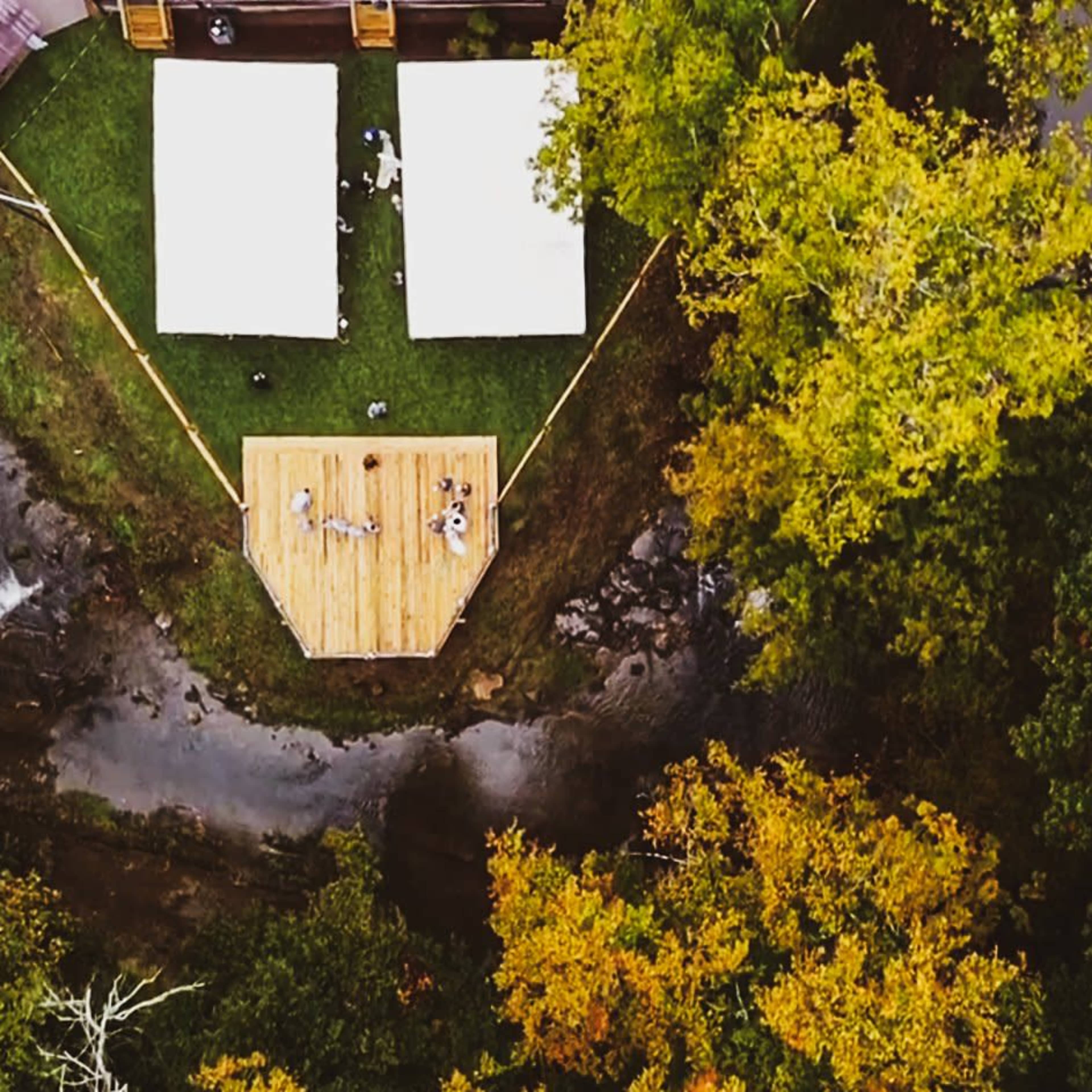 An aerial view shows a wooden platform surrounded by tents and trees in a forested area with a creek nearby.