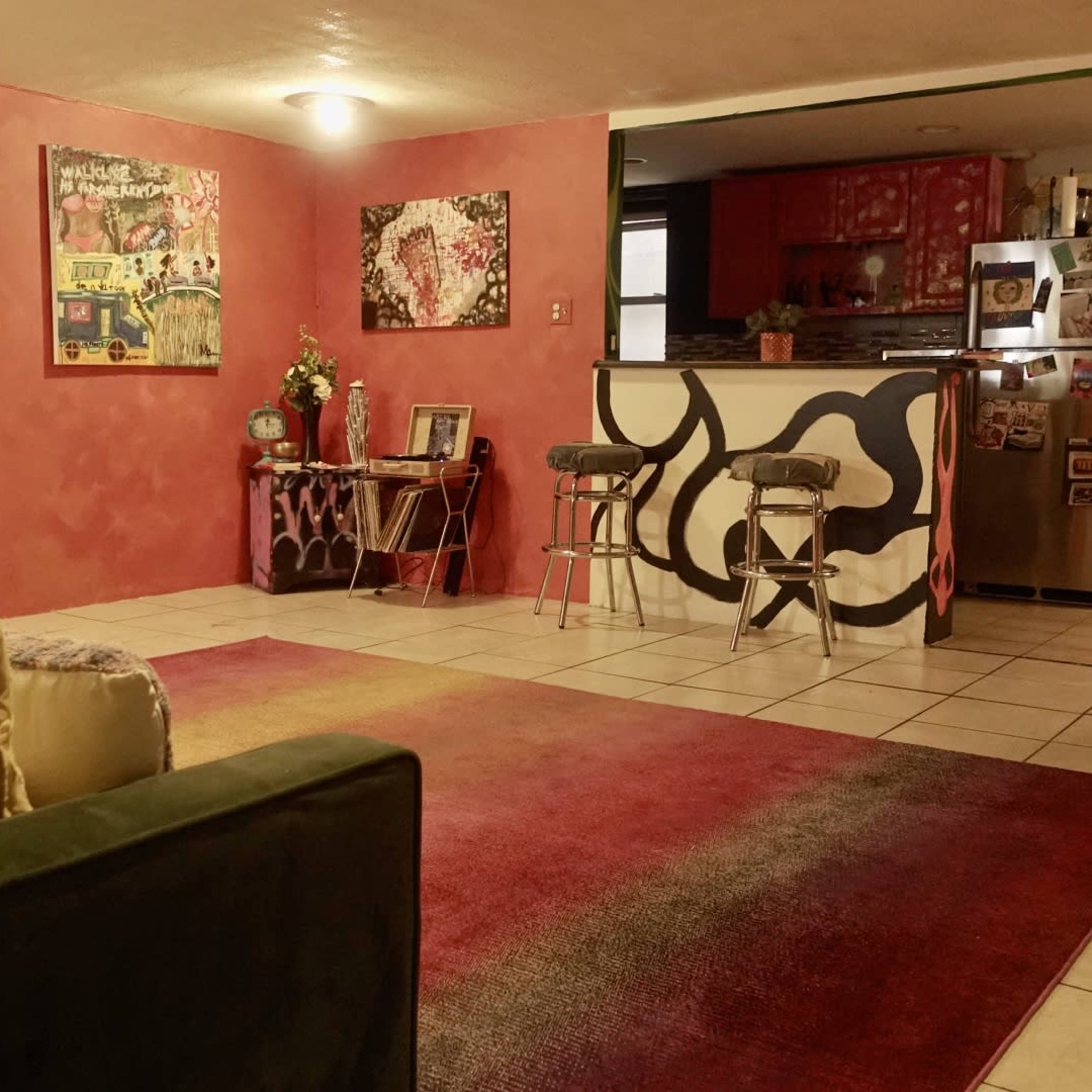 A brightly colored living room features a large area rug, eclectic artwork on the walls, and a kitchen visible in the background with a bar counter and stools.