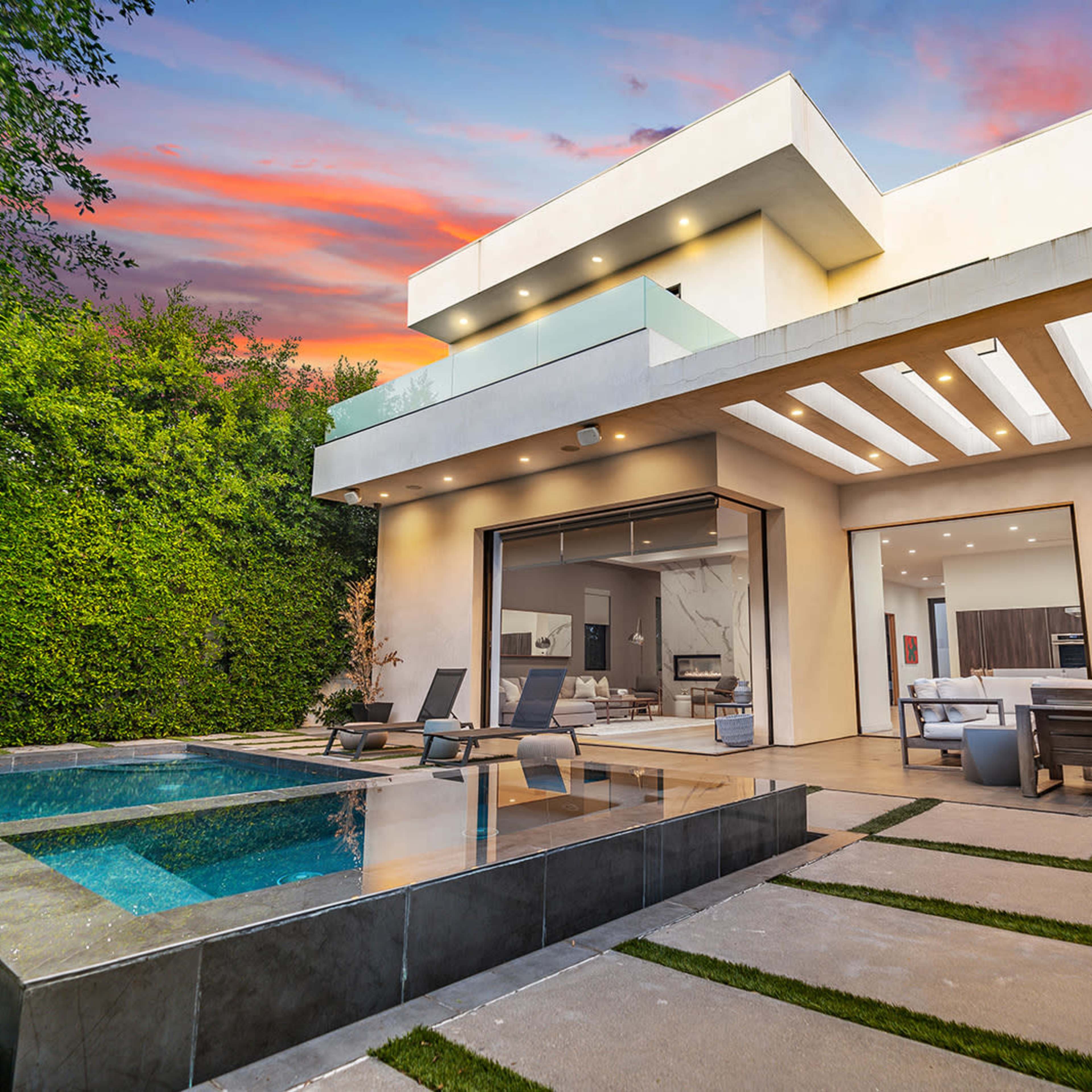 A modern house with a pool is set against a colorful sunset and surrounded by greenery.