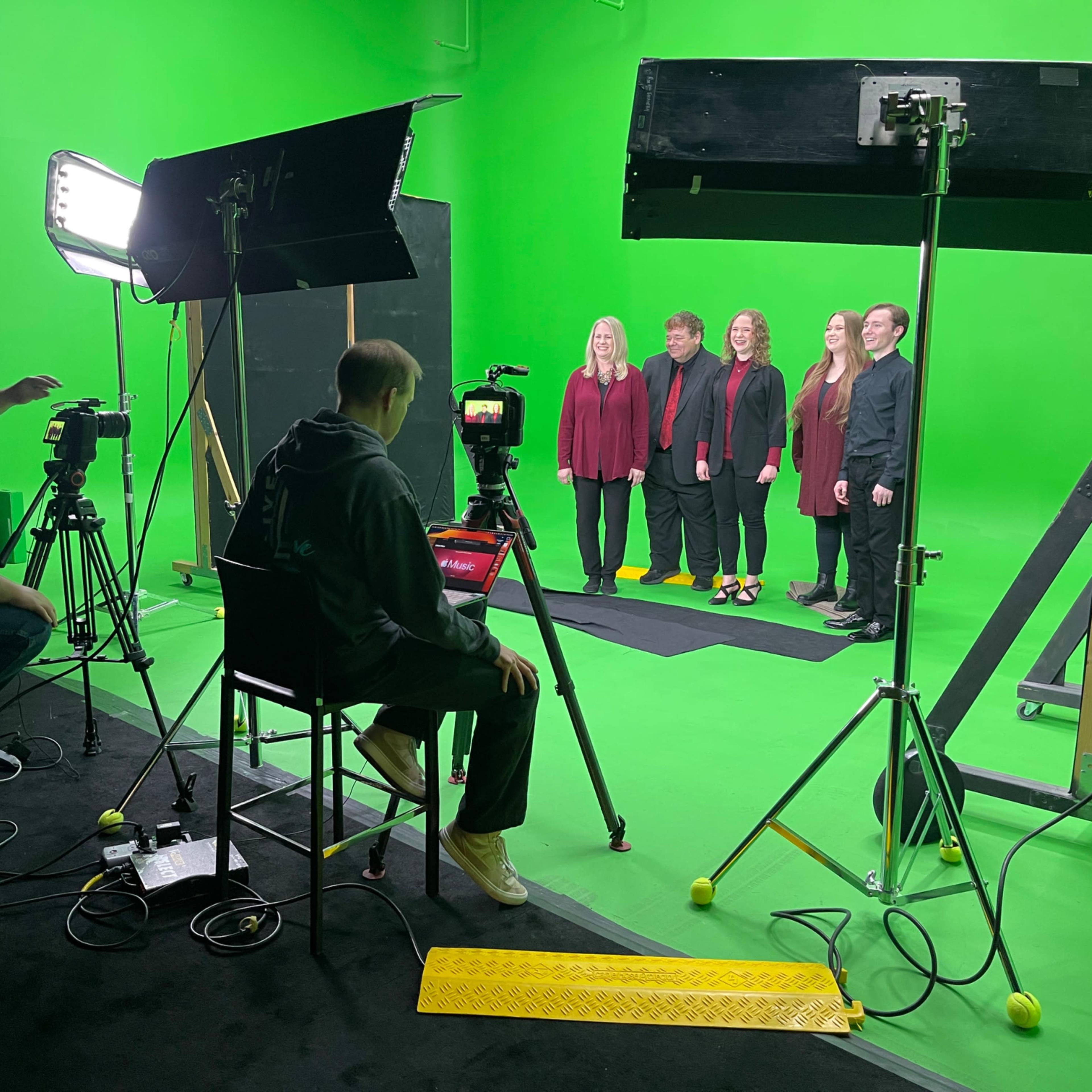 A group of five people stands in front of a green screen while two camera operators and a lighting setup are positioned nearby.