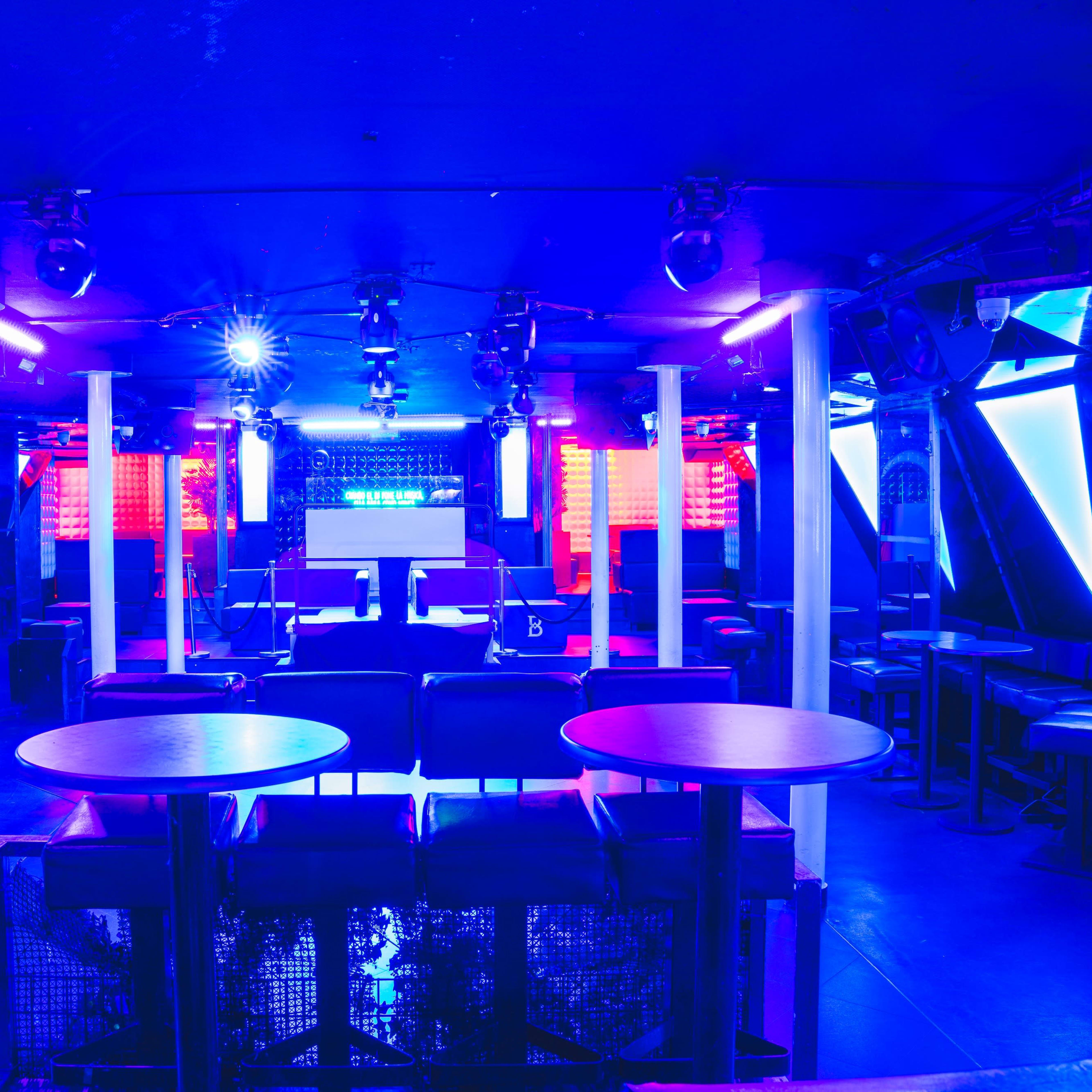 A dimly lit nightclub with blue lighting, empty tables, and a DJ booth in the background.