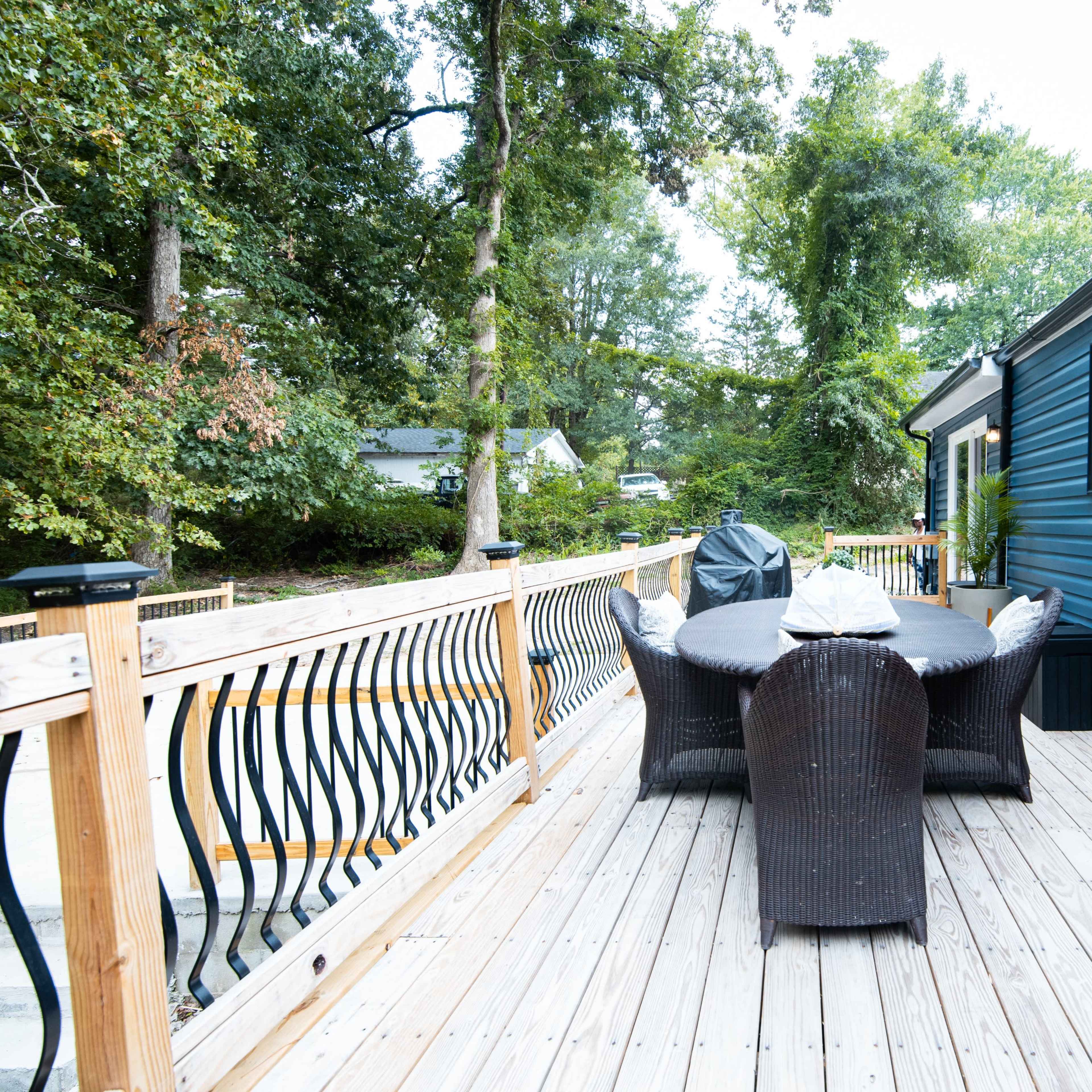 A wooden deck with a table and six chairs, surrounded by trees and a house featuring a blue exterior wall.