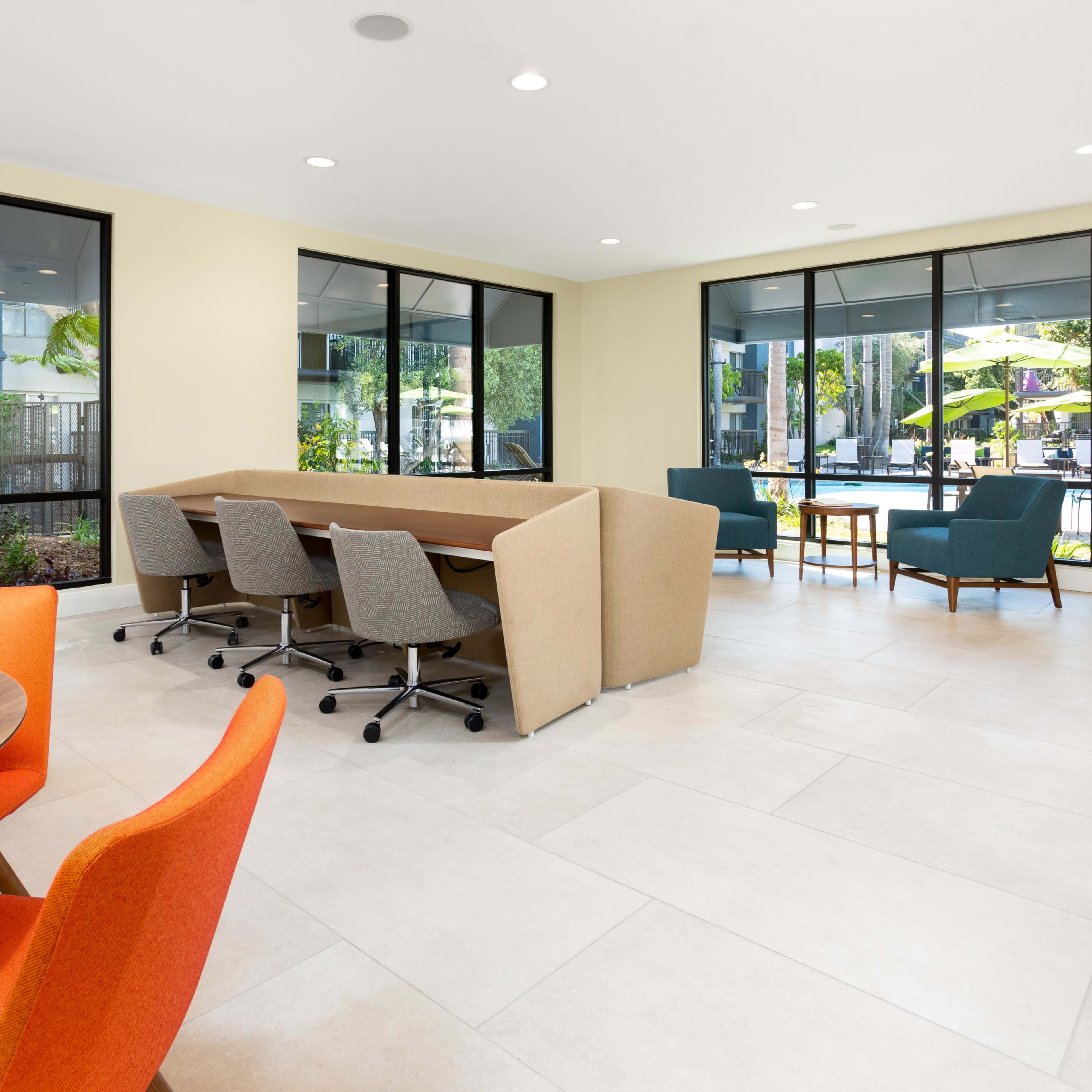 A modern community lounge area with round and rectangular tables, colorful chairs, and large windows overlooking an outdoor space.