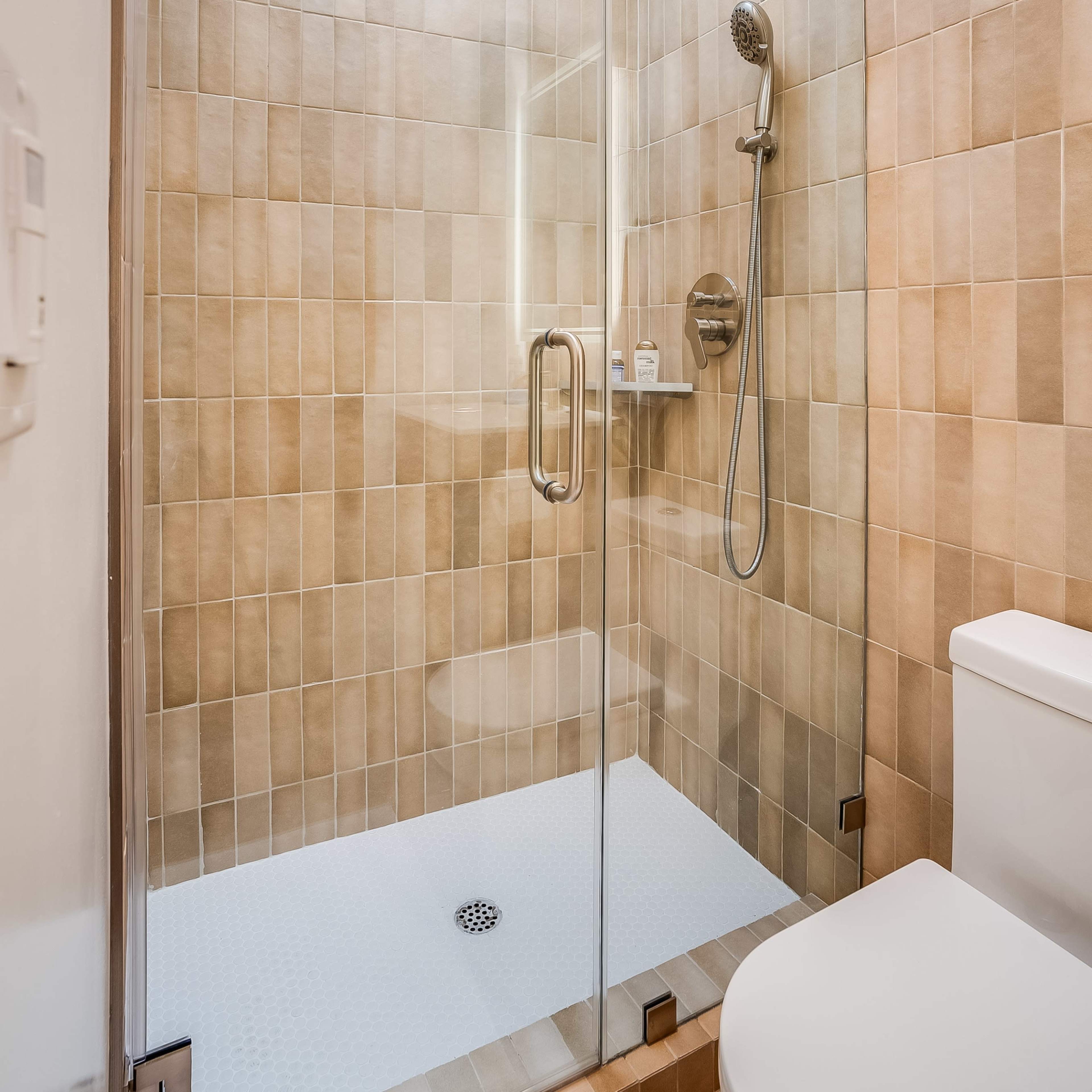 A modern glass-enclosed shower with a tiled wall and a white toilet in a small bathroom.