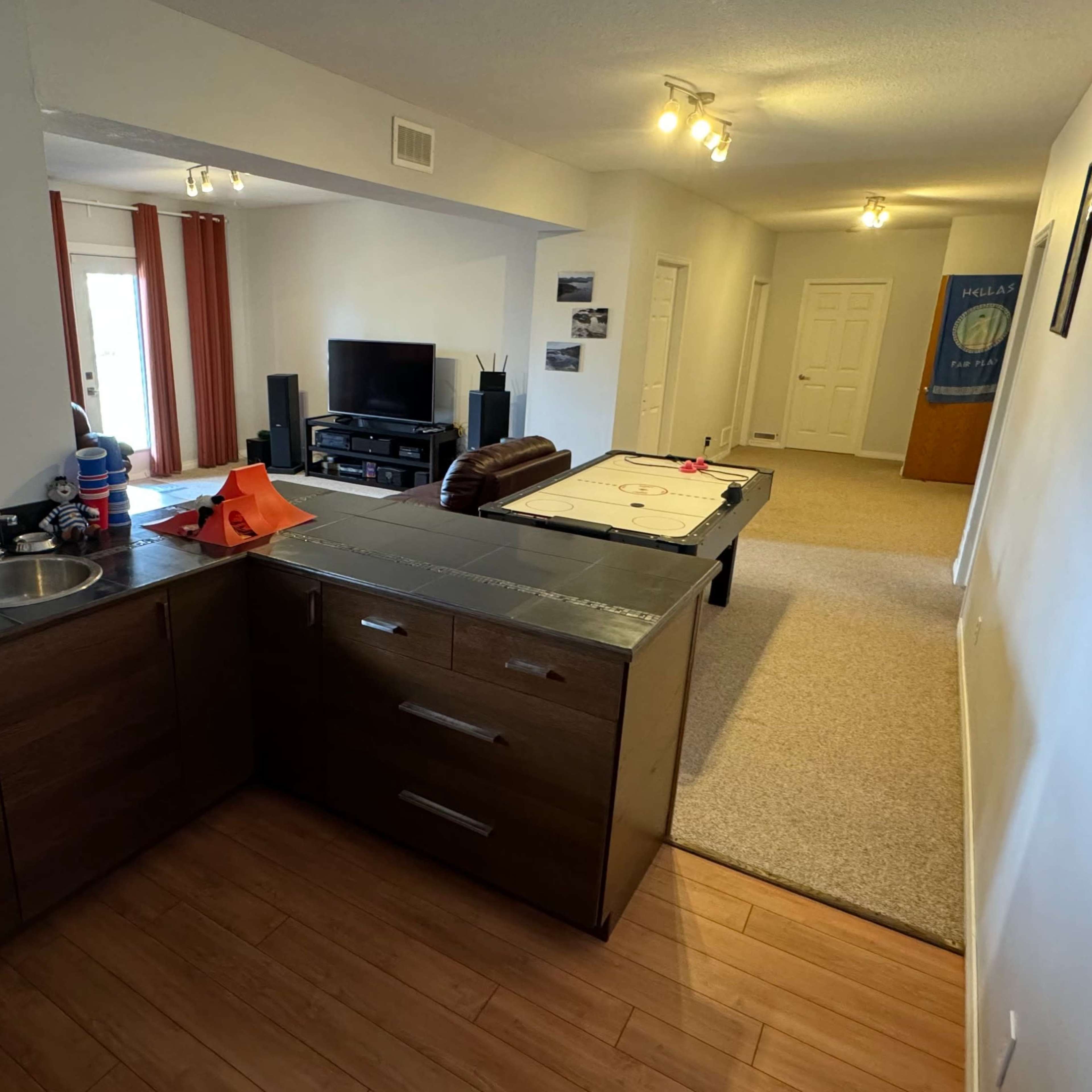 An open-plan living area with a kitchen on the left and a living room area featuring a TV and a pool table in the background.