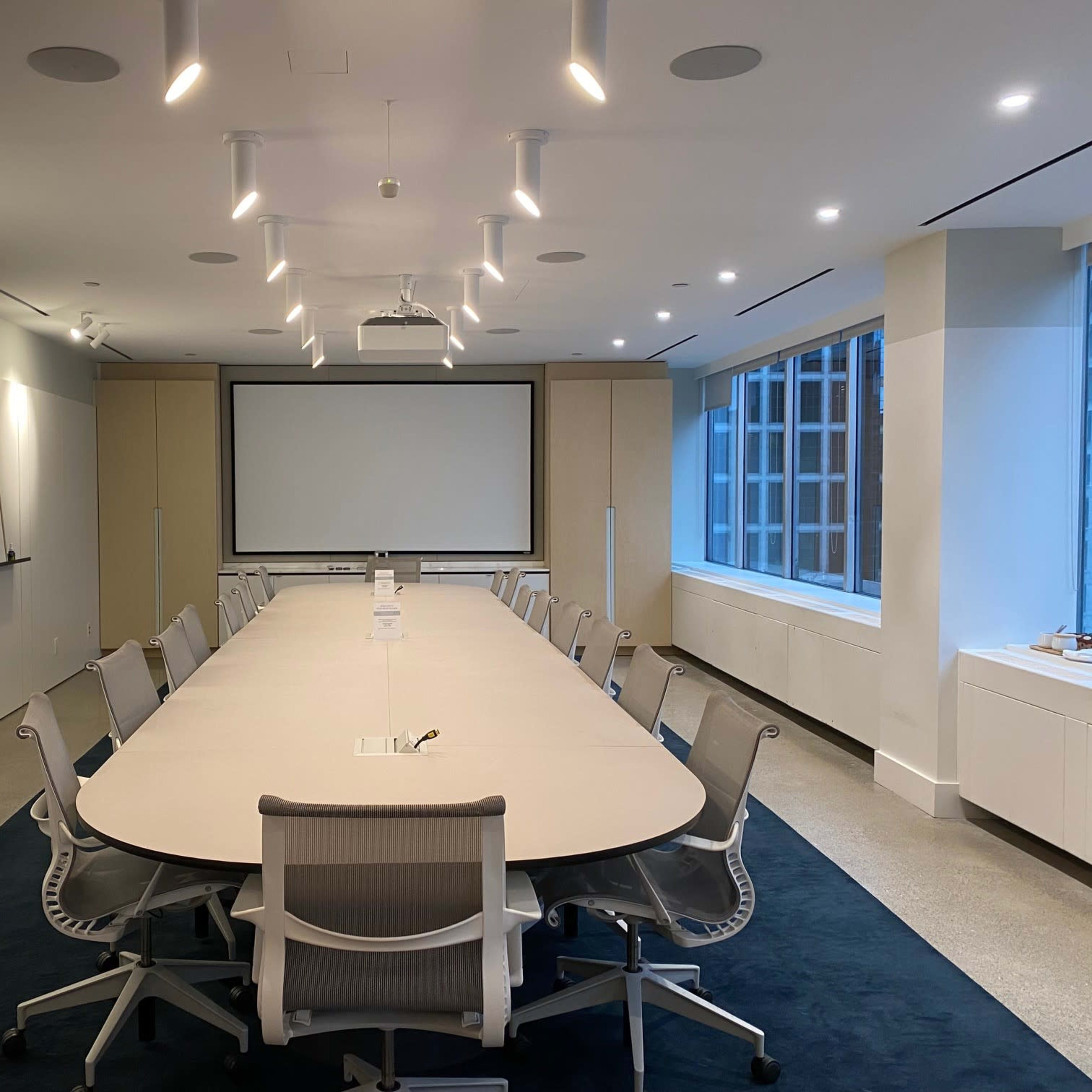 A large, modern conference room features a long oval table surrounded by ergonomic chairs, with a projector screen at one end and whiteboards on the walls.