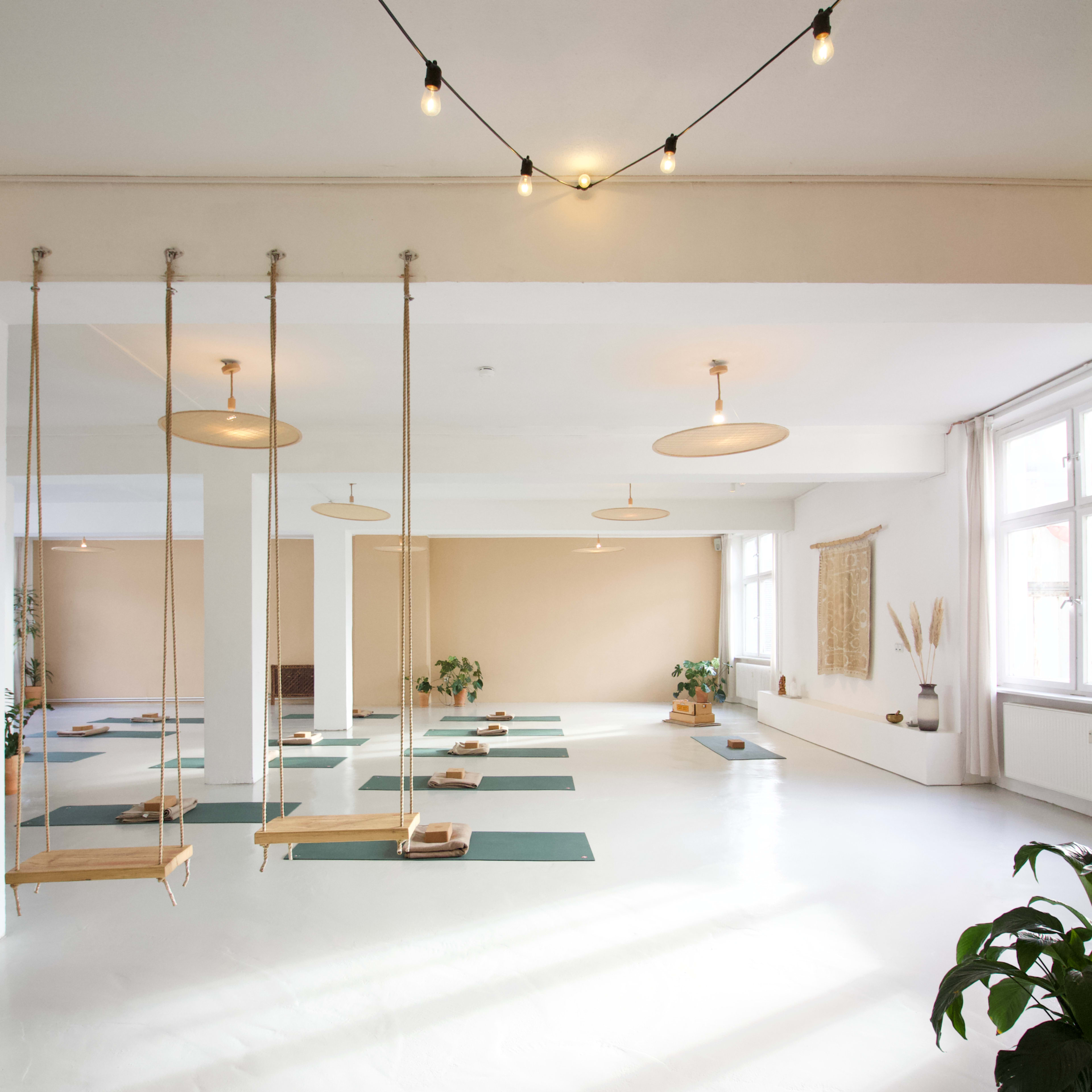 A spacious, well-lit room with yoga mats arranged on the floor and swings hanging from the ceiling.