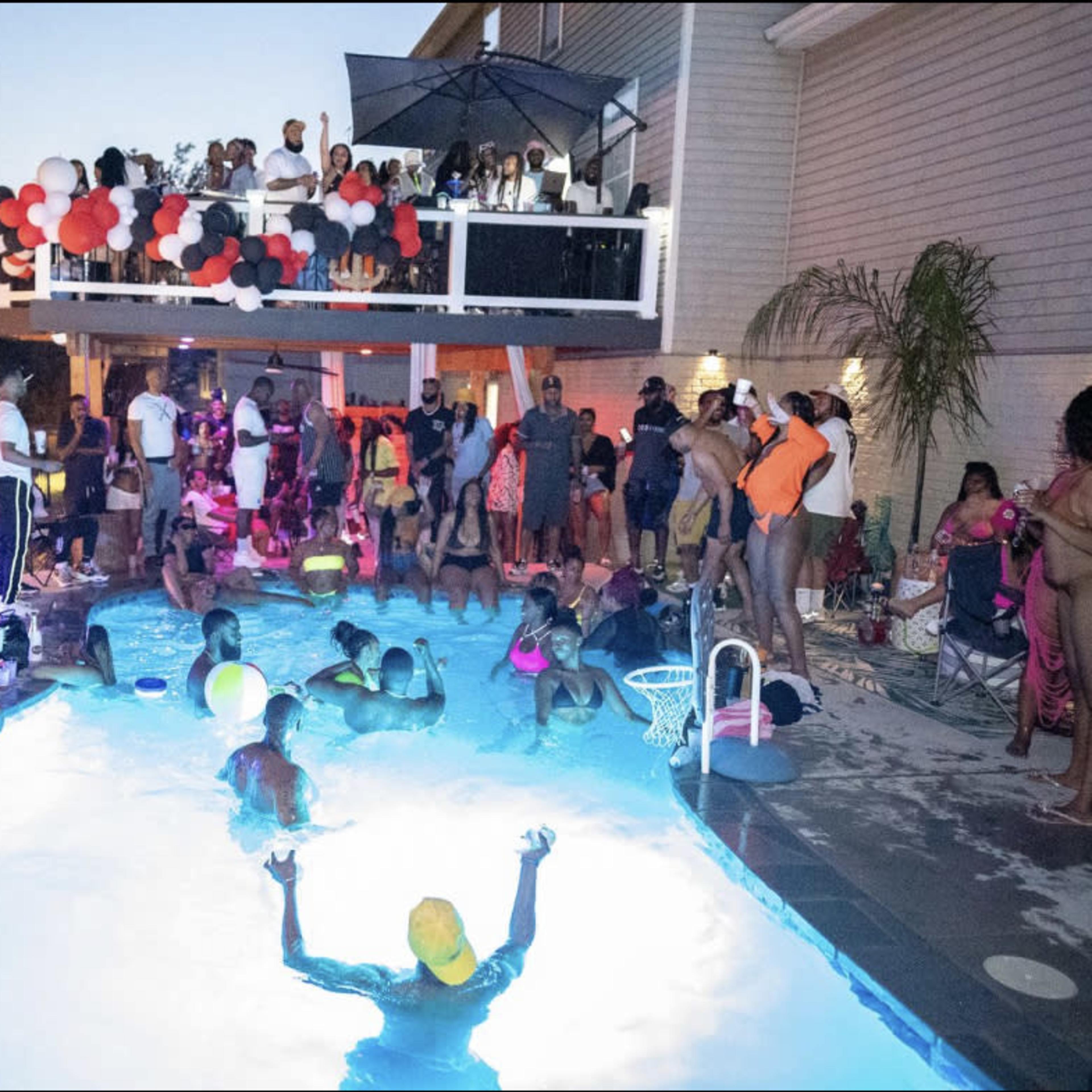 A large group of people gathers around a pool at twilight, with some in the water and others on the deck, enjoying a lively party atmosphere.