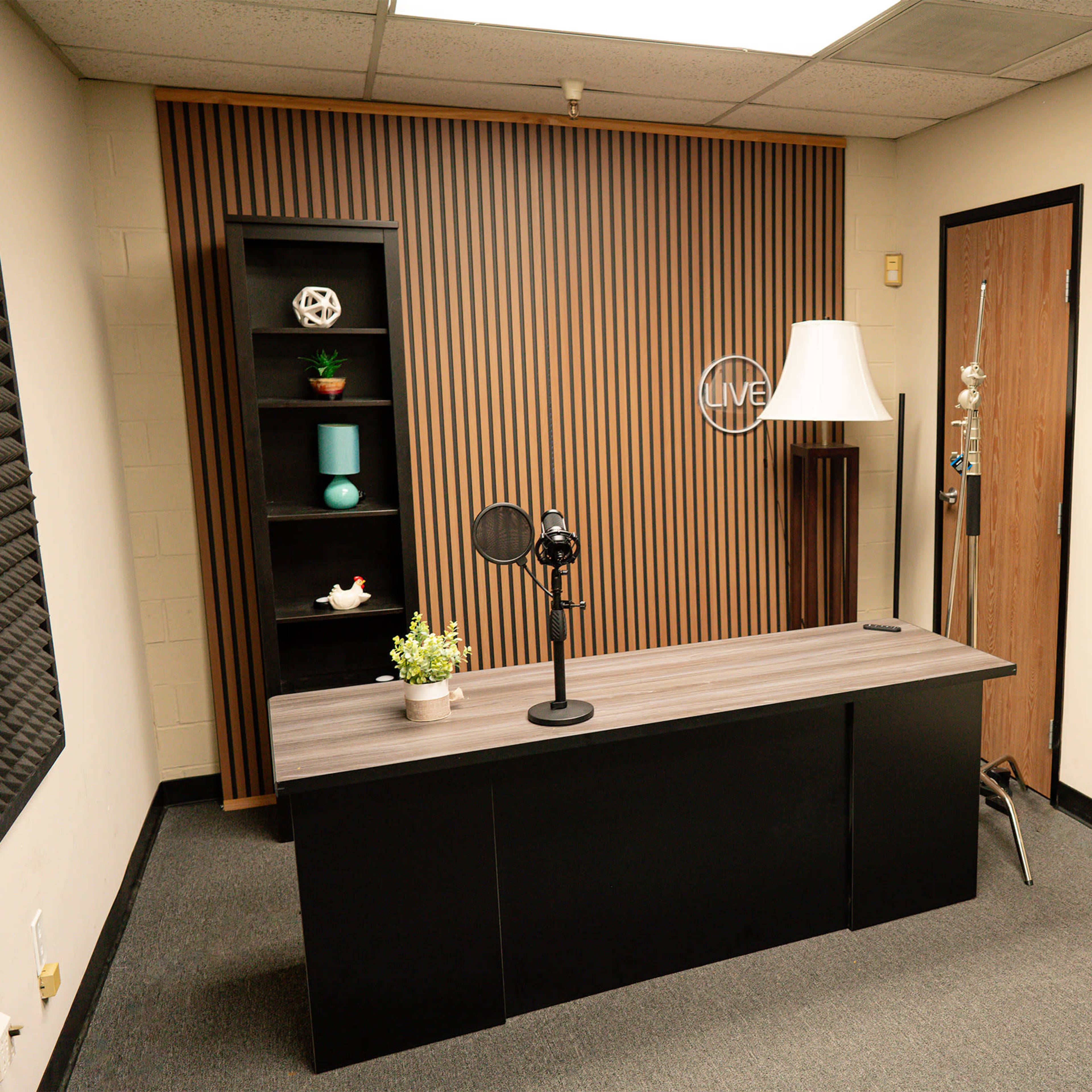 A small recording studio with a desk, a microphone, soundproof panels on the walls, and decorative items on a shelf.