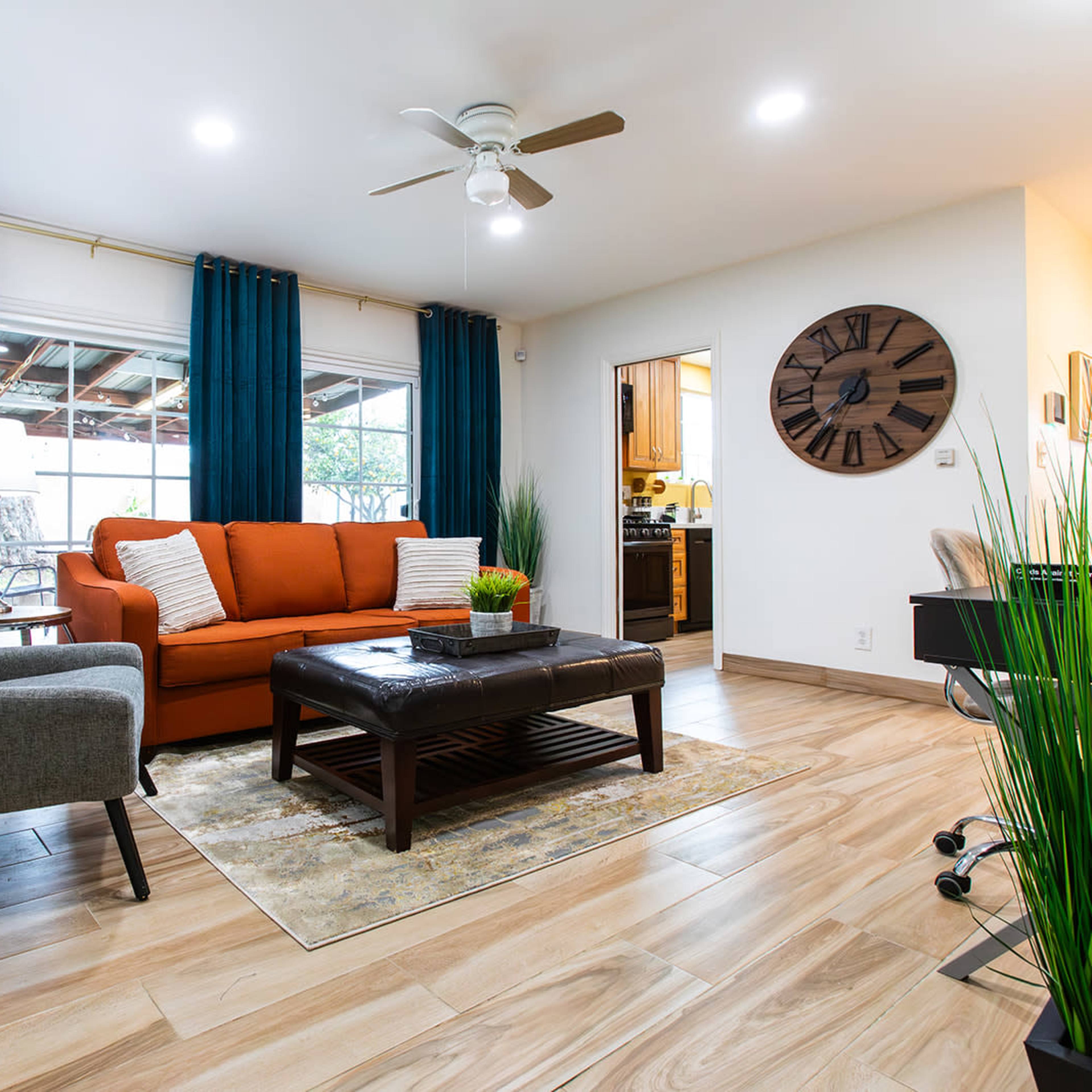 A living room featuring an orange sofa, a gray armchair, a coffee table on a patterned rug, and potted plants, with a kitchen visible in the background.