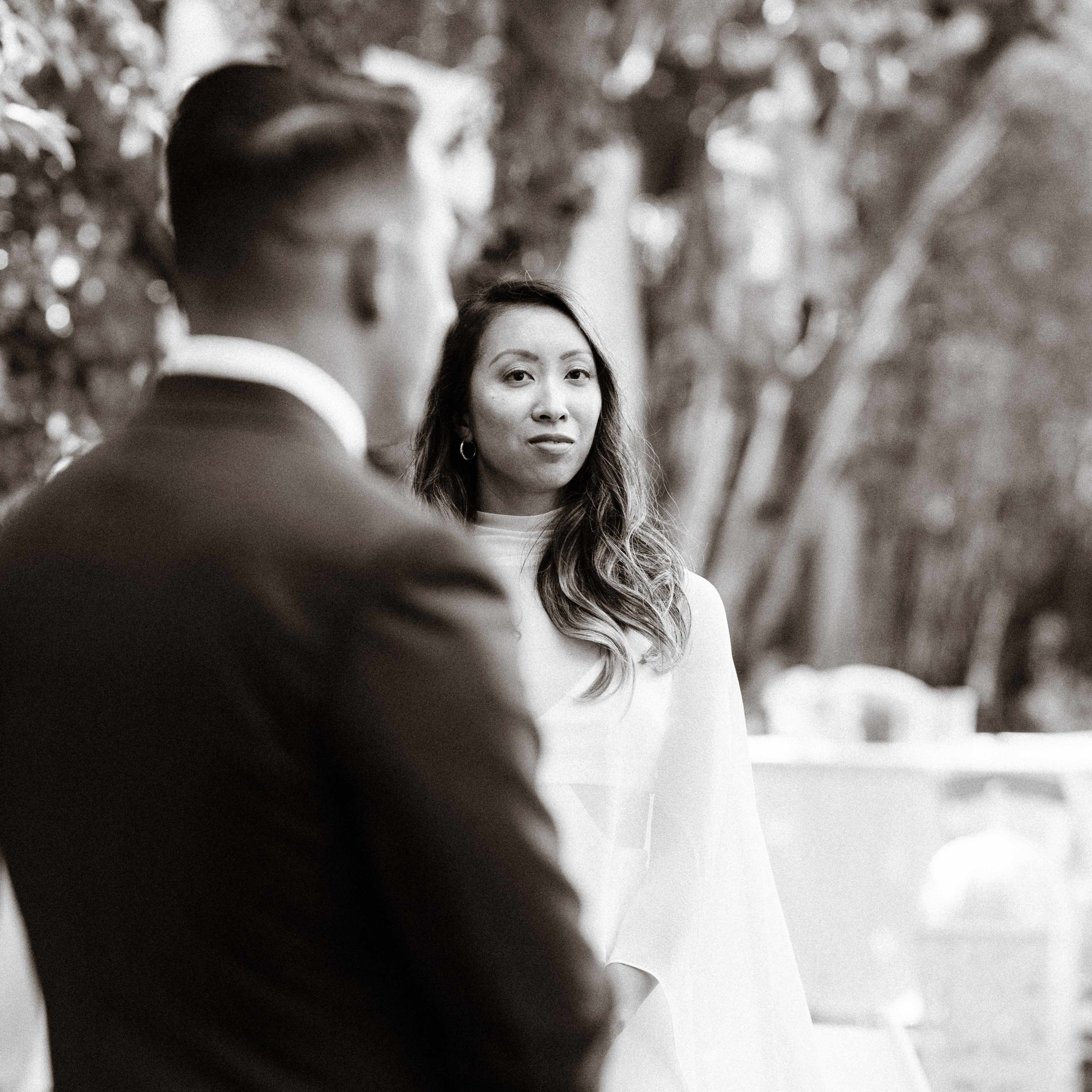 A man in a suit stands facing a woman dressed in white at an outdoor setting surrounded by trees.