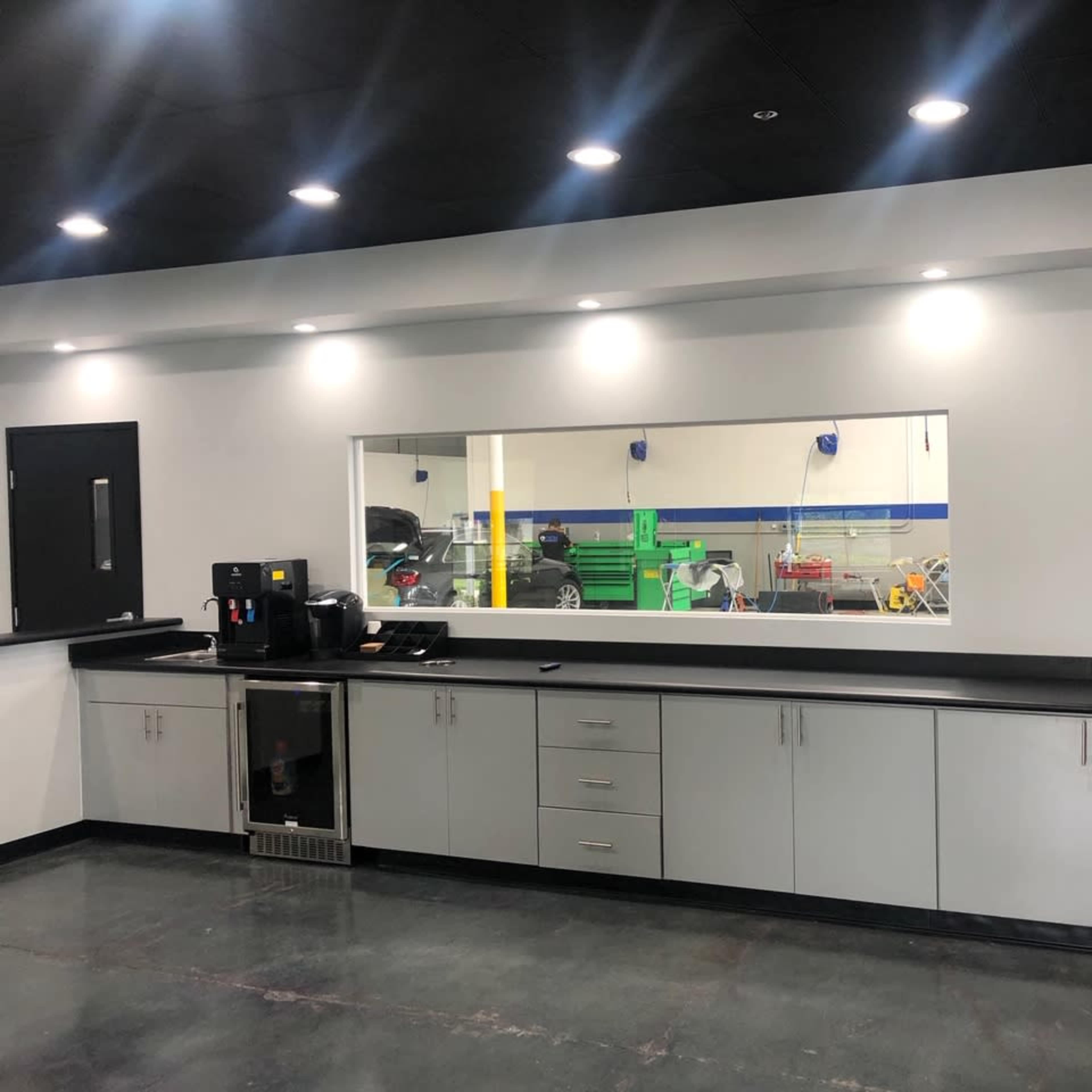 A modern kitchen area with a long countertop, a coffee machine, a small refrigerator, and a large window revealing a workspace behind it.